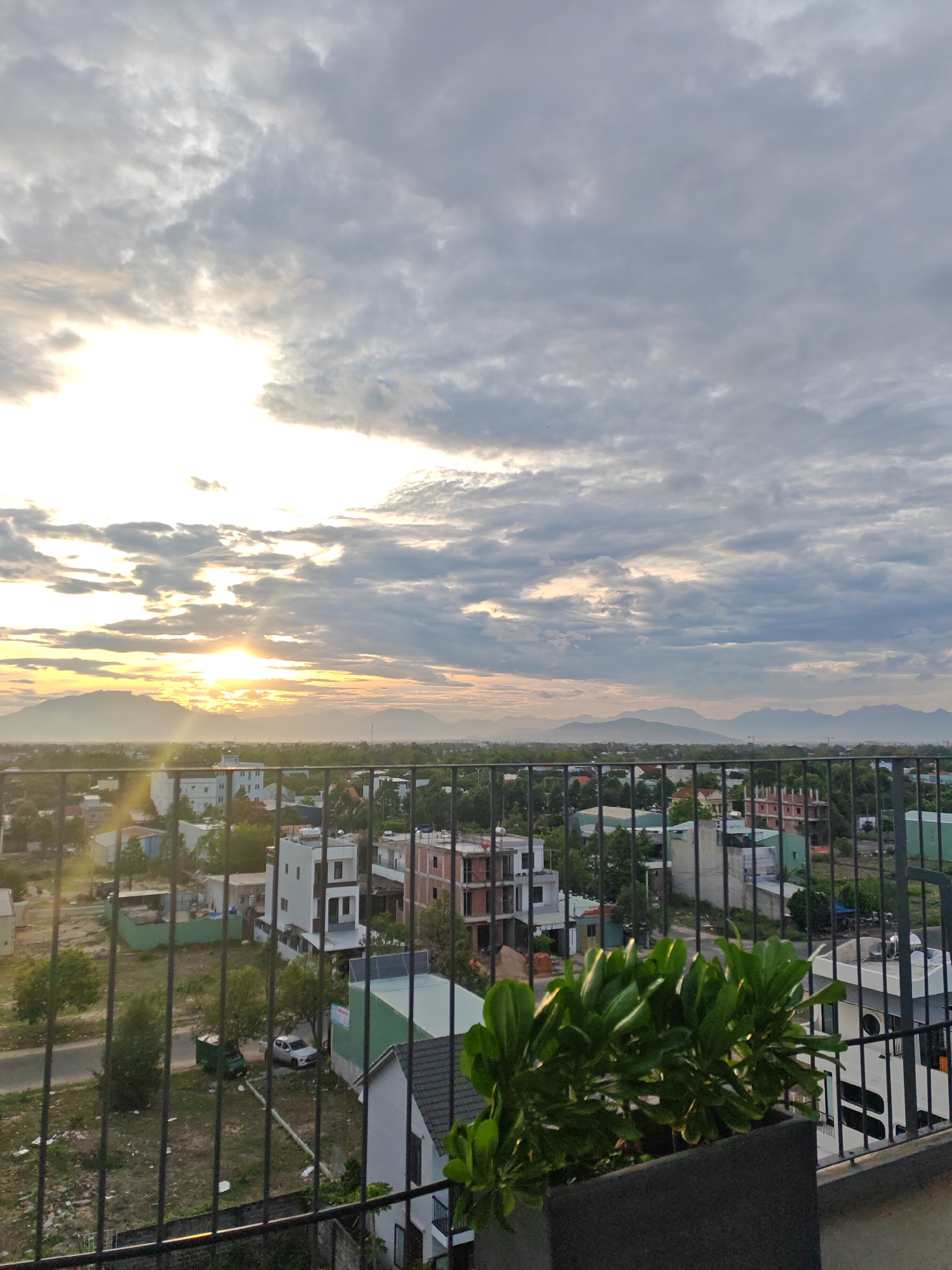This image depicts a panoramic view of a cityscape, likely Đà Nẵng, Vietnam, during sunset. The sun, a bright orb, is partially obscured by dramatic, cloudy skies, casting a warm, golden light across the horizon. In the foreground, a dark metal railing creates a boundary, with lush green plants in a dark planter box positioned to the right. Beyond the railing, the scene unfolds with a cluster of buildings, a mix of modern multi-story structures and smaller houses, interspersed with trees and vegetation. In the mid-ground, a road is visible with a parked silver SUV and a small green vehicle. The background is dominated by a range of distant mountains, their silhouettes softened by the atmospheric haze. The overall impression is one of urban development coexisting with natural landscapes under a striking sky.
