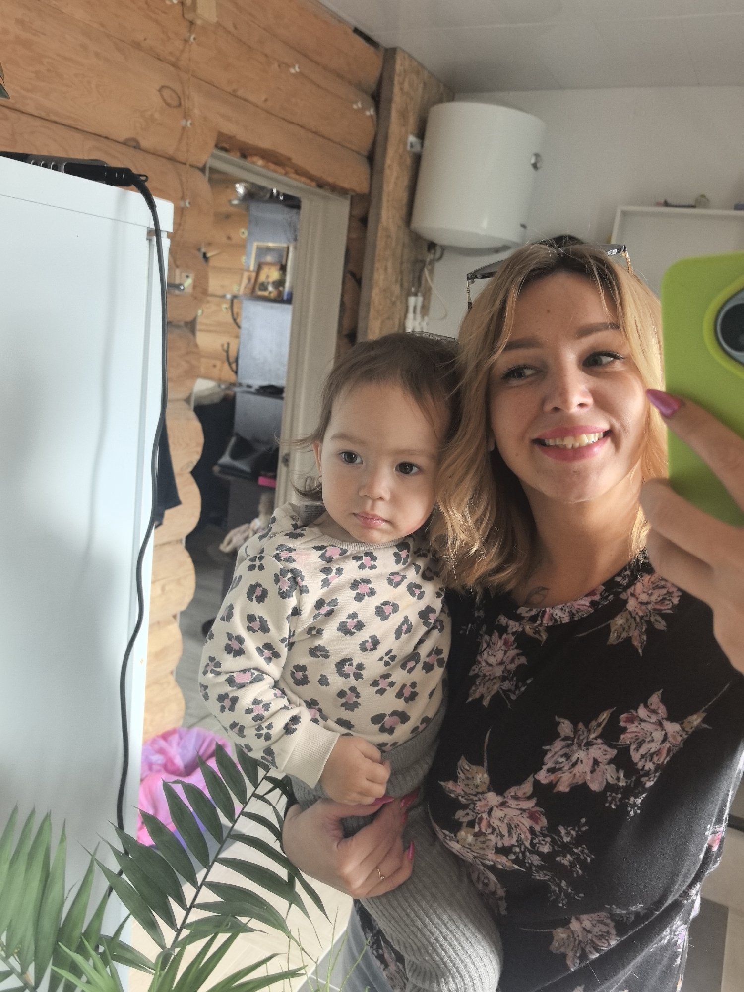 A woman holds a toddler in a selfie taken indoors, likely in Noviy, Russia, based on the wooden log cabin style of the interior walls. The woman has blonde hair and is wearing a black floral print top. The toddler has brown hair and is wearing a beige patterned sweatshirt. The woman is holding a green phone. In the background is a white water heater and a doorway leading to another room. A large white appliance is to the left. A potted plant sits in the foreground.