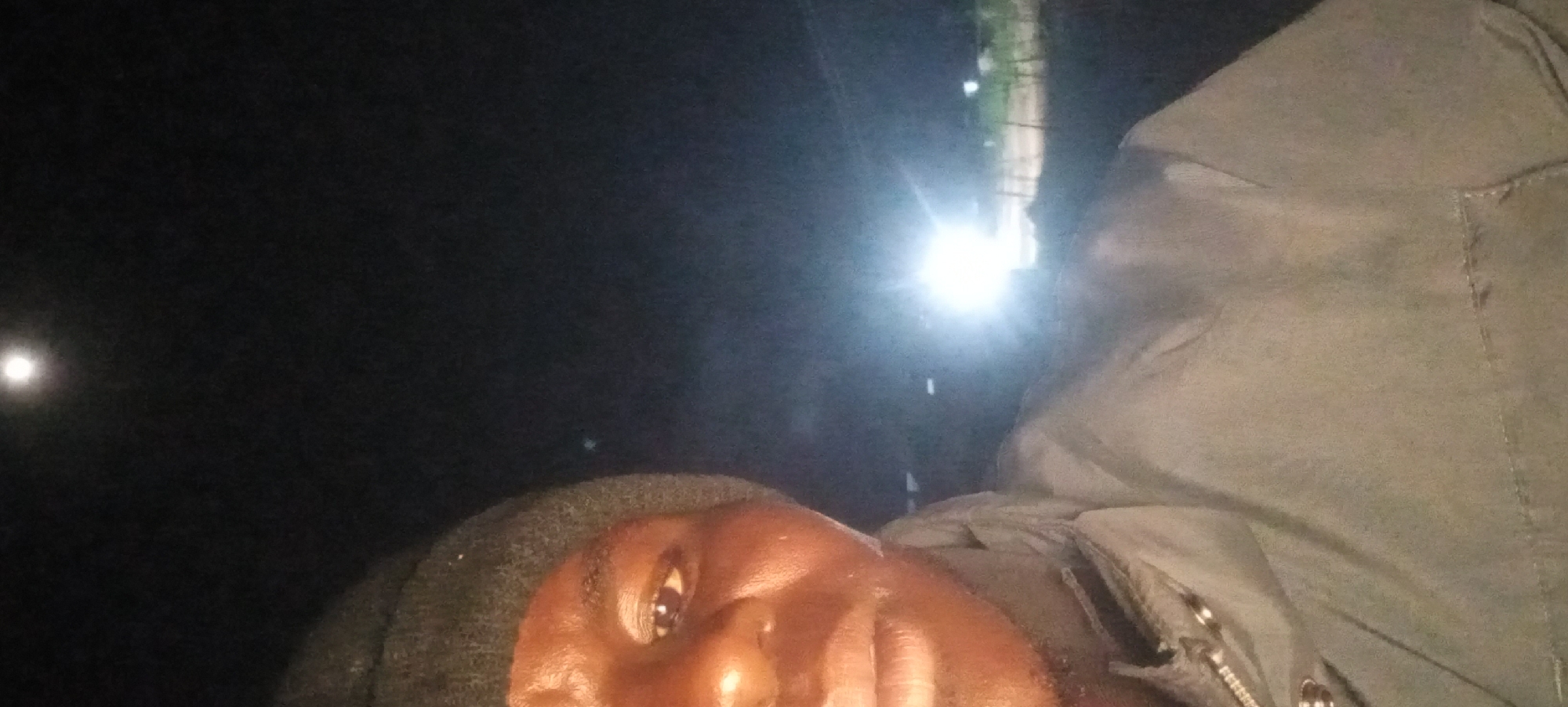 The image is a selfie taken at night in Kwang, Nigeria. The subject is a person wearing a dark beanie and a light-colored jacket. They are looking towards the camera, with part of their face and upper torso visible. In the background, there is a bright light source, likely from a vehicle or streetlights, illuminating the scene. The sky above is dark, with a faint, bright circle that could be the moon or a distant light. The environment appears to be outdoors, possibly on a street or in a rural area, with some vegetation visible on the right side of the image.