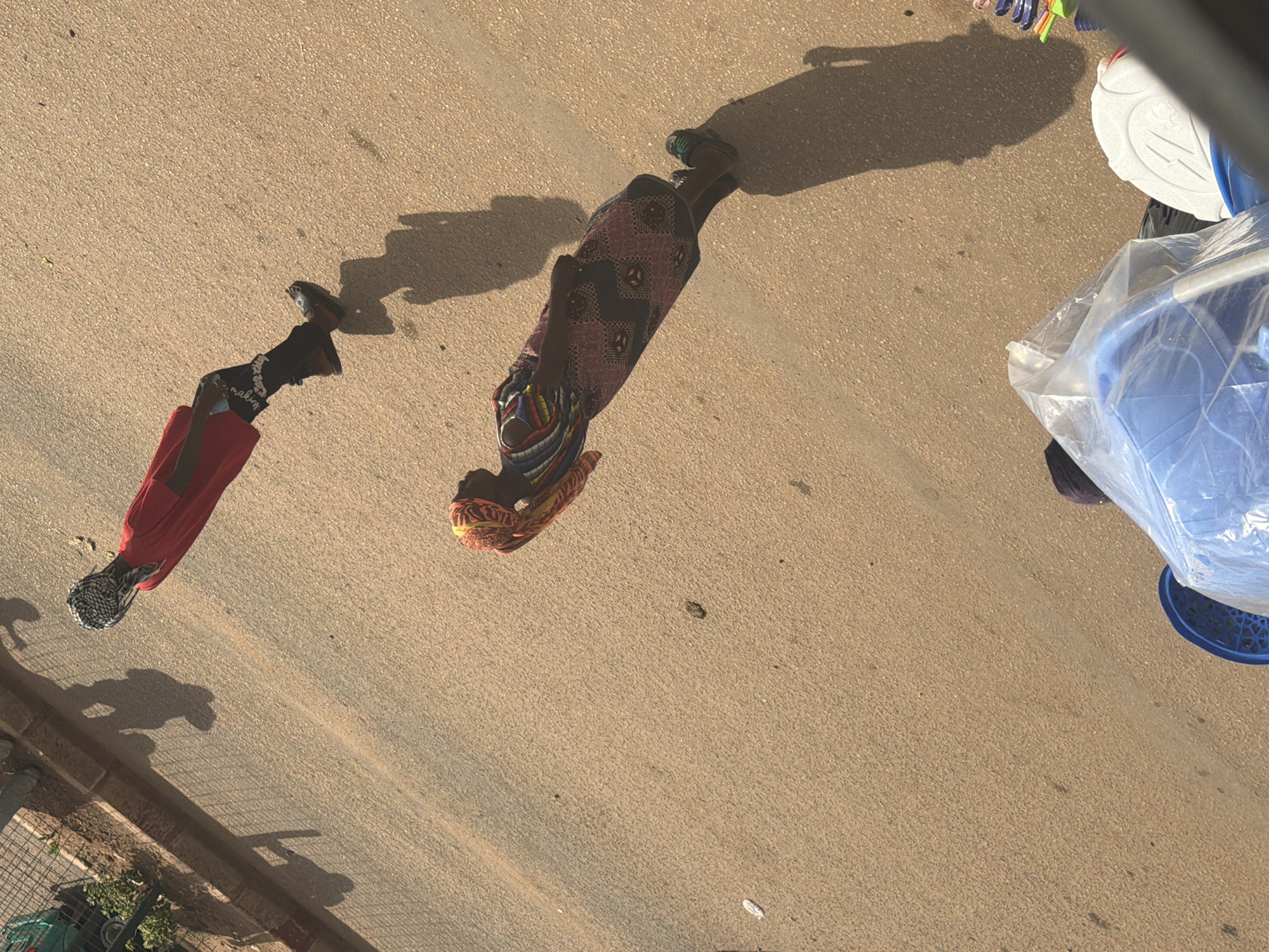The image captures two individuals walking on a paved surface, likely a road in Bukuru, Nigeria. The scene is outdoors, and the strong shadows cast on the ground suggest it is daytime, possibly late afternoon. One person, closer to the left of the frame, wears a long, flowing red garment and a patterned headscarf. Their legs are visible, with dark shoes or sandals. The other individual, further to the right, is adorned in a patterned dress with vibrant colors and a matching headscarf. Their legs are also visible, wearing patterned sandals.
In the upper right portion of the image, several objects are visible, including a white container with an embossed design, possibly a cooler or storage bin. Next to it is a large blue object partially covered in clear plastic sheeting, and a blue plastic basket. These objects, along with the paved surface and the surrounding sparse vegetation hinted at on the lower left, suggest an informal market or roadside setting.
The activity depicted is simple: the two individuals are walking, their paths converging slightly but not interacting. The overall impression is one of everyday life in a Nigerian community. There are no discernible emotions conveyed by the subjects, as their faces are not clearly visible or are obscured by head coverings. The lighting and the presence of shadows are the primary cues for the time of day.