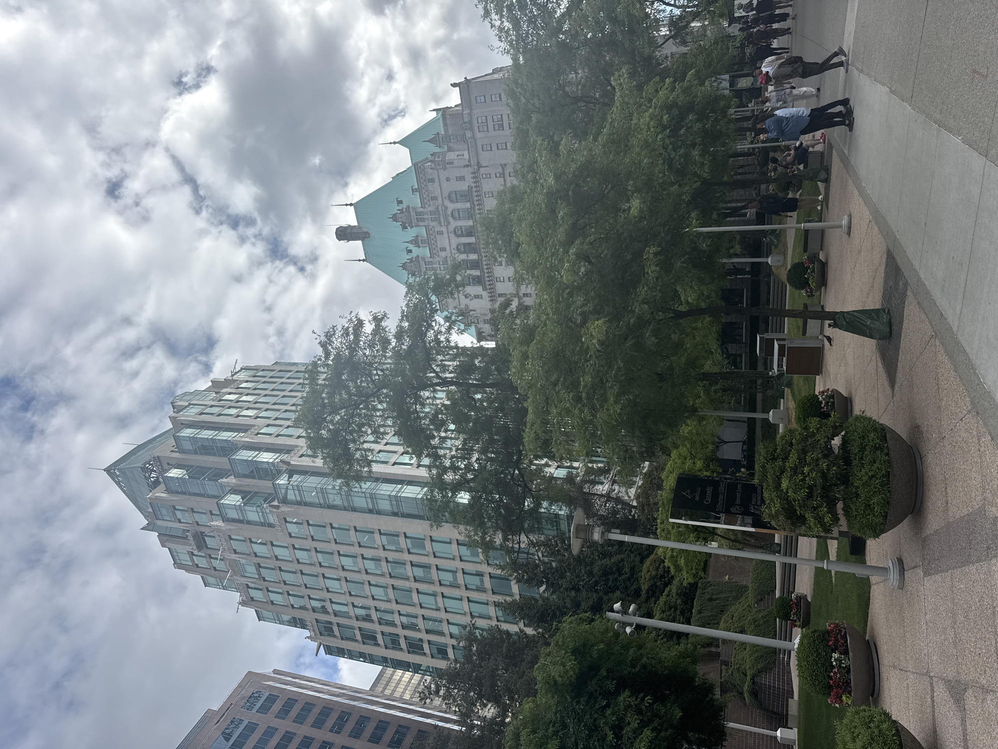 This is a wide-angle, eye-level shot of a city street scene in Vancouver, Canada, on a cloudy day. In the background, two prominent buildings are visible: a modern, glass-facade skyscraper with a pointed, green roof, and to its right, a more ornate, older building with a distinctive turquoise roof and castle-like architectural details. The older building appears to be the Fairmont Empress Hotel.
In the foreground, a paved walkway lines the right side of the image, with a grassy area and planter boxes with decorative shrubs and flowers to the left. Several people are walking on the sidewalk and standing in small groups, dressed in casual clothing. A street lamp and a dark trash bin are also present in the mid-ground. Lush green trees are scattered throughout the scene, providing shade and separating the buildings from the street level. The overall impression is one of an urban environment with a mix of historical and modern architecture, enhanced by landscaping and the presence of pedestrians.