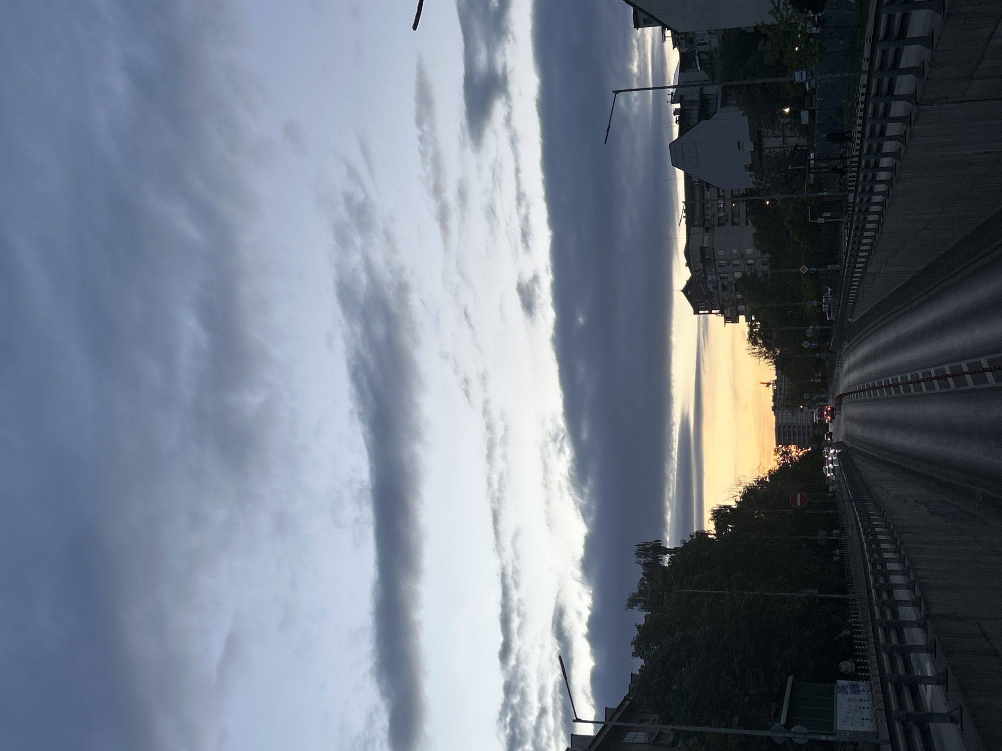 The image shows a street in Sofia, Bulgaria, facing east at sunset. A wide, grey roadway is visible, flanked by concrete barriers with black and white stripes. Buildings line the sides of the street, partially obscured by trees. A line of traffic is visible in the distance, with illuminated headlights. The sky is dominated by long, striated clouds, ranging in color from white to dark grey, with the sunset visible as a streak of orange light. Streetlights are visible along the roadway.