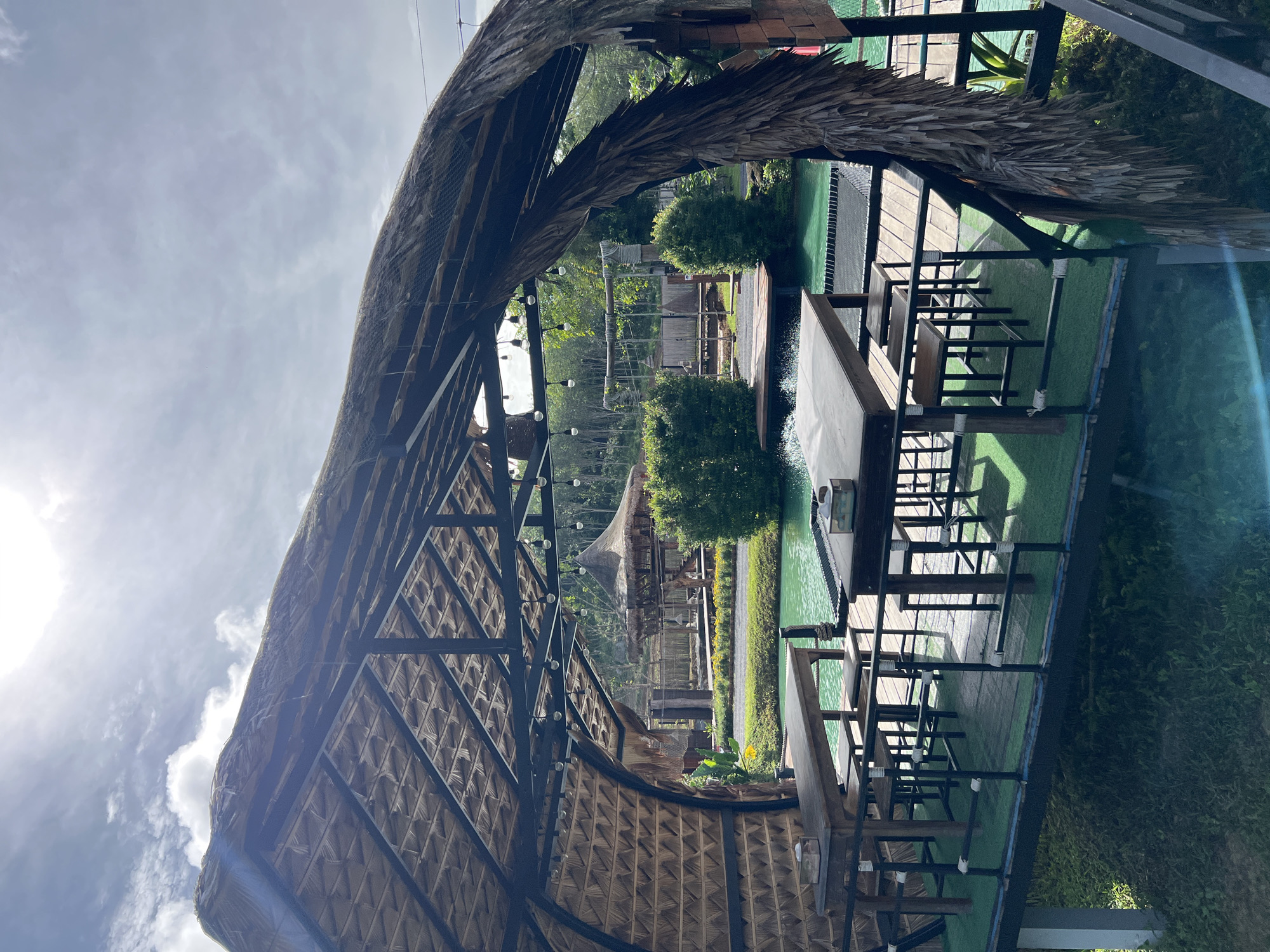 This is an outdoor scene, likely a restaurant or dining area, situated in a tropical environment. The main subjects are the various structures and furniture designed for patrons. A prominent feature is a thatched roof, supported by a metal frame, which extends over a seating area. Underneath this roof, several wooden tables and chairs are arranged on a raised platform with a green, artificial turf-like surface.
Beyond this immediate seating area, a vibrant green body of water is visible, possibly a pool or a pond. Another thatched-roof structure, smaller than the first, can be seen in the background, nestled amongst lush green trees. The landscape suggests a natural, possibly jungle-like setting with abundant vegetation. The overall impression is one of a tranquil, open-air dining experience.
The time of day appears to be daytime, with bright sunlight casting shadows and creating highlights, particularly a noticeable lens flare on the right side of the image. The sky is partly cloudy, with a mix of bright sunlight and diffused light. No people are visible in the frame, and there is no visible text. The location cues, such as the abundant greenery and the style of architecture, align with a tropical setting like Tambon Pa Kae Bo Hin, Thailand.