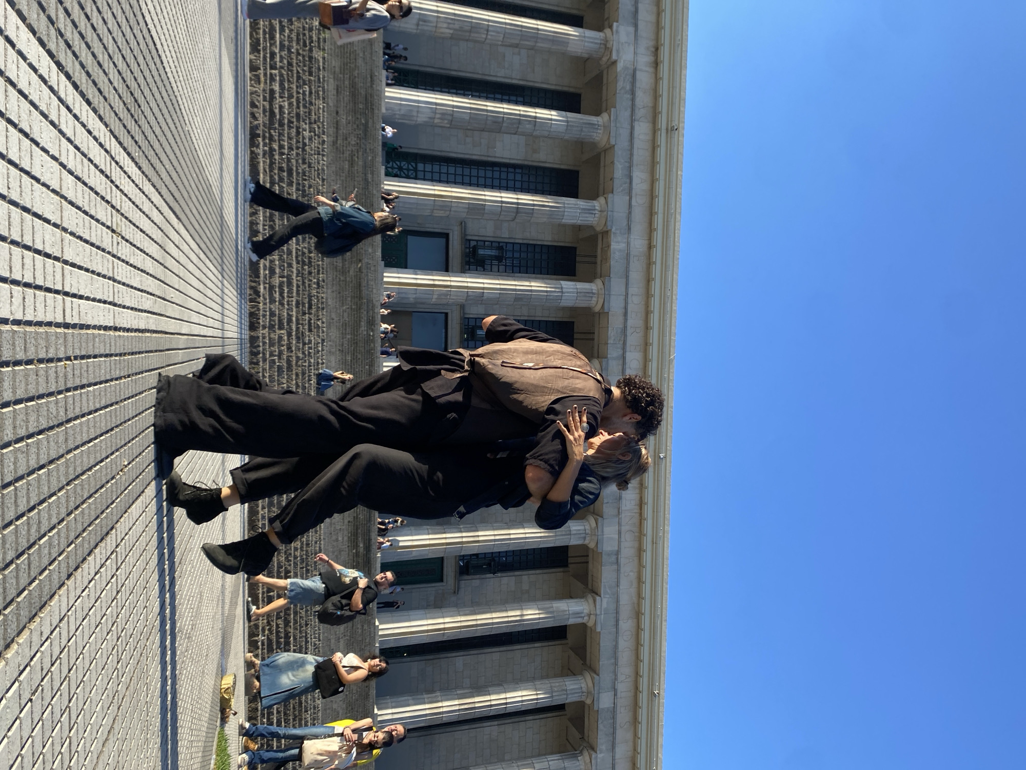 The image shows a couple kissing in front of the Facultad de Derecho of the University of Buenos Aires in Buenos Aires, Argentina. The man is wearing black pants and a black shirt with a brown backpack. The woman is also wearing black pants and a black shirt. Other people are walking in the background. The building is large, with multiple pillars, and the sky is clear and blue. The ground is paved with rows of light gray bricks.