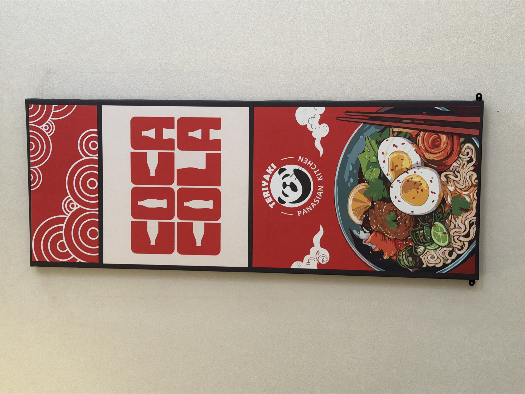 The image shows a tall, narrow vertical sign mounted on a textured beige wall. The sign is divided into three horizontal sections, each with a distinct design.
The top section features a red background with a pattern of concentric white circles, resembling stylized clouds or ripples.
The middle section is a white rectangle with large, red, block letters spelling "COCA COLA". The letters are stylized, with the "C"s containing an outline of a Coca-Cola bottle, and the "A"s also incorporating bottle shapes.
The bottom section is a vibrant red square containing a circular logo and an illustration of a bowl of food. The logo at the top of this section depicts a black and white panda's face within a circle. Text around the panda reads "TERIYAKI" above and "PANASIAN KITCHEN" below, with stylized white clouds on either side. Below the logo is an overhead view of a bowl of ramen. The ramen consists of noodles, broth, sliced hard-boiled eggs with visible yolks, slices of lime, pieces of meat or tofu, and leafy greens, all in a dark blue-grey bowl. Two red chopsticks rest diagonally against the right side of the bowl.
The sign appears to be a promotional display for a restaurant or food establishment. There are no people visible in the image. The lighting is even, suggesting it is indoors. The wall has a subtle, uniform texture. The sign is held in place by small black brackets at the bottom corners.
Based on the visual elements, particularly the "Panasian Kitchen" branding combined with the illustration of ramen and the panda logo, this sign is likely located at a restaurant named "Teriyaki Panasian Kitchen" in Tashkent, Uzbekistan. The prevalence of such establishments and their typical decorative signage supports this location.