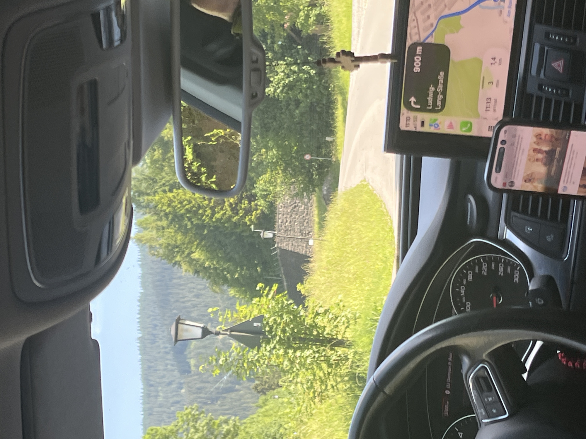 The image shows the interior of a car, looking out the windshield and through the rearview mirror. The car is on a paved road that curves to the right, surrounded by lush green trees and hills. A street lamp is visible on the left side of the road. The dashboard displays a navigation system showing a route to "Ludwig-Lang-Straße" in an urban area, indicating the car is likely in or near a city. The GPS shows a distance of 900 meters to the destination. A phone is mounted on the dashboard, displaying what appears to be a social media feed or web page. The time on the navigation system is 11:13 AM, with an estimated arrival time in 3 minutes, covering a distance of 1.4 km. The overall environment suggests a sunny day in a scenic, possibly mountainous region, with the car in motion. Given the navigation details and the architecture visible (stone walls, traditional street lamps), it is consistent with a location in a European town.