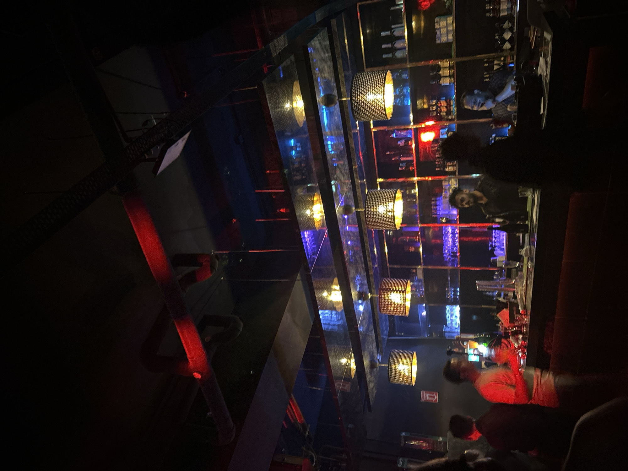 The image captures a dimly lit bar in Kuala Lumpur, Malaysia. Several people are present, including bartenders and patrons. One bartender is visible behind the bar, interacting with customers. The bar itself is well-stocked with bottles and glasses. A series of light fixtures hang above the bar, illuminating the scene. A red pipe adds another visual element to the image.