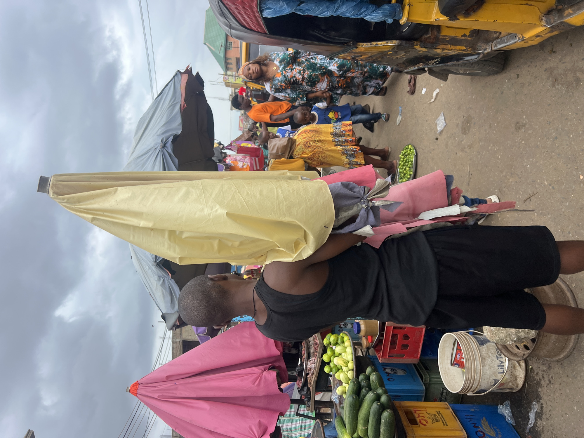 The image depicts a bustling outdoor market scene in Nkpor, Nigeria, under a cloudy sky. A man in a black tank top and shorts is holding a large, folded yellow umbrella over his left shoulder. To his left, a bright pink umbrella is visible. In the foreground, a pile of cucumbers sits in blue crates. A metal plate filled with small green fruits, likely limes or small tomatoes, is also present.
Further into the market, several people are seen. A woman in a vibrantly patterned dress, her hair wrapped in a patterned scarf, walks with a young boy wearing a blue shirt. Another woman in a yellow dress with an orange pattern walks ahead of them. In the background, a man in an orange shirt stands near a yellow auto-rickshaw, commonly known as a Keke napep. Stalls are set up with various goods, including what appears to be meat on a platter behind the pink umbrella. Crates and buckets are scattered throughout the market, indicating commercial activity. The ground is paved with asphalt or concrete and appears wet in places, suggesting recent rain or a wet market environment. The overall atmosphere is one of lively commerce and everyday life in a Nigerian urban setting.