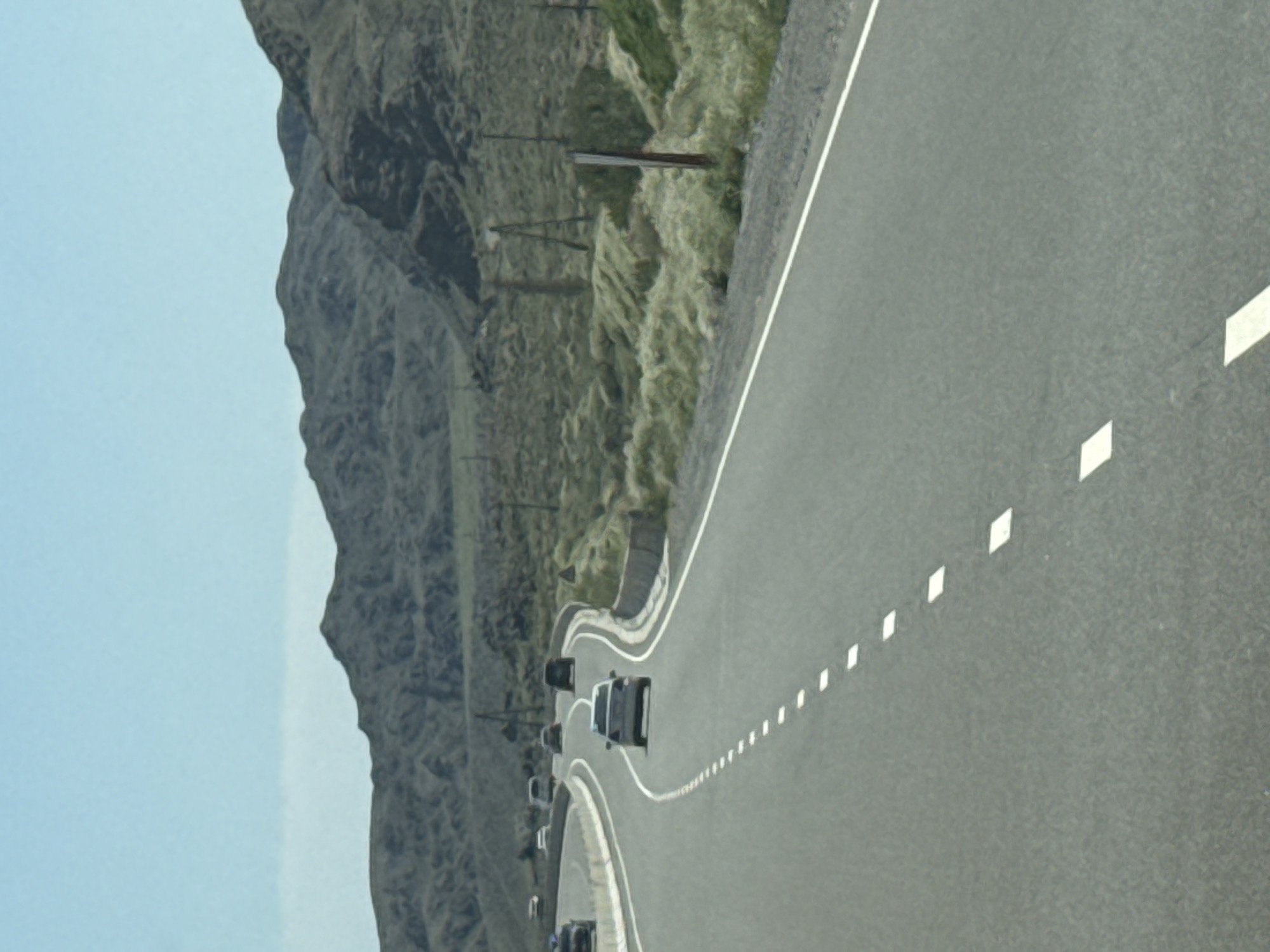 The image depicts a highway scene in the vicinity of Kok Moynok Eki, Kyrgyzstan. A grey asphalt road curves through a landscape of arid, rolling hills and mountains under a clear blue sky. Several cars are visible on the road: a silver sedan is in the foreground, driving away from the viewer, followed by other vehicles further down the highway. The roadside is characterized by dry, sparse vegetation and scattered wooden posts, suggesting a remote, undeveloped area. The overall impression is one of travel through a mountainous, desert-like environment.