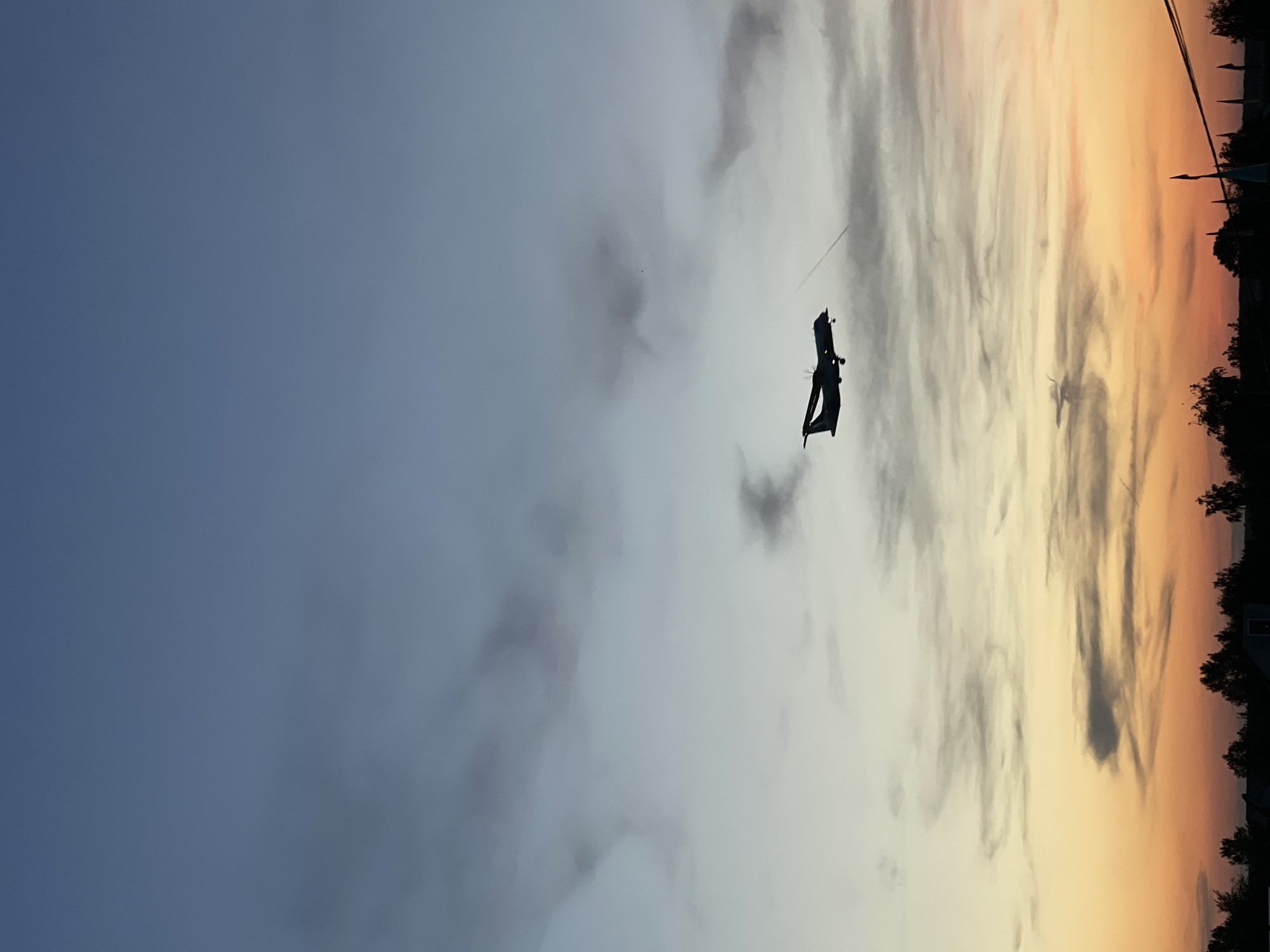 The image displays a twilight sky, likely at sunset, with a prominent dark silhouette of a flying object. The sky exhibits a clear gradient, transitioning from a deep blue-gray on the left to lighter grays and then to warm orange and yellow tones towards the right. Scattered clouds are present, some appearing dark against the lighter sky, others catching the golden light.
The main subject is an angular, dark object, possibly a large kite or a specialized drone, flying in the upper mid-right section of the frame. A thin line, consistent with a kite string or tether, extends upwards from it. Along the far right edge of the image, a dark, silhouetted horizon line reveals the tops of trees and several distant structures, suggesting a town or residential area. No people are visible, and there is no discernible text within the image. The scene conveys a calm and atmospheric moment during dusk or dawn.