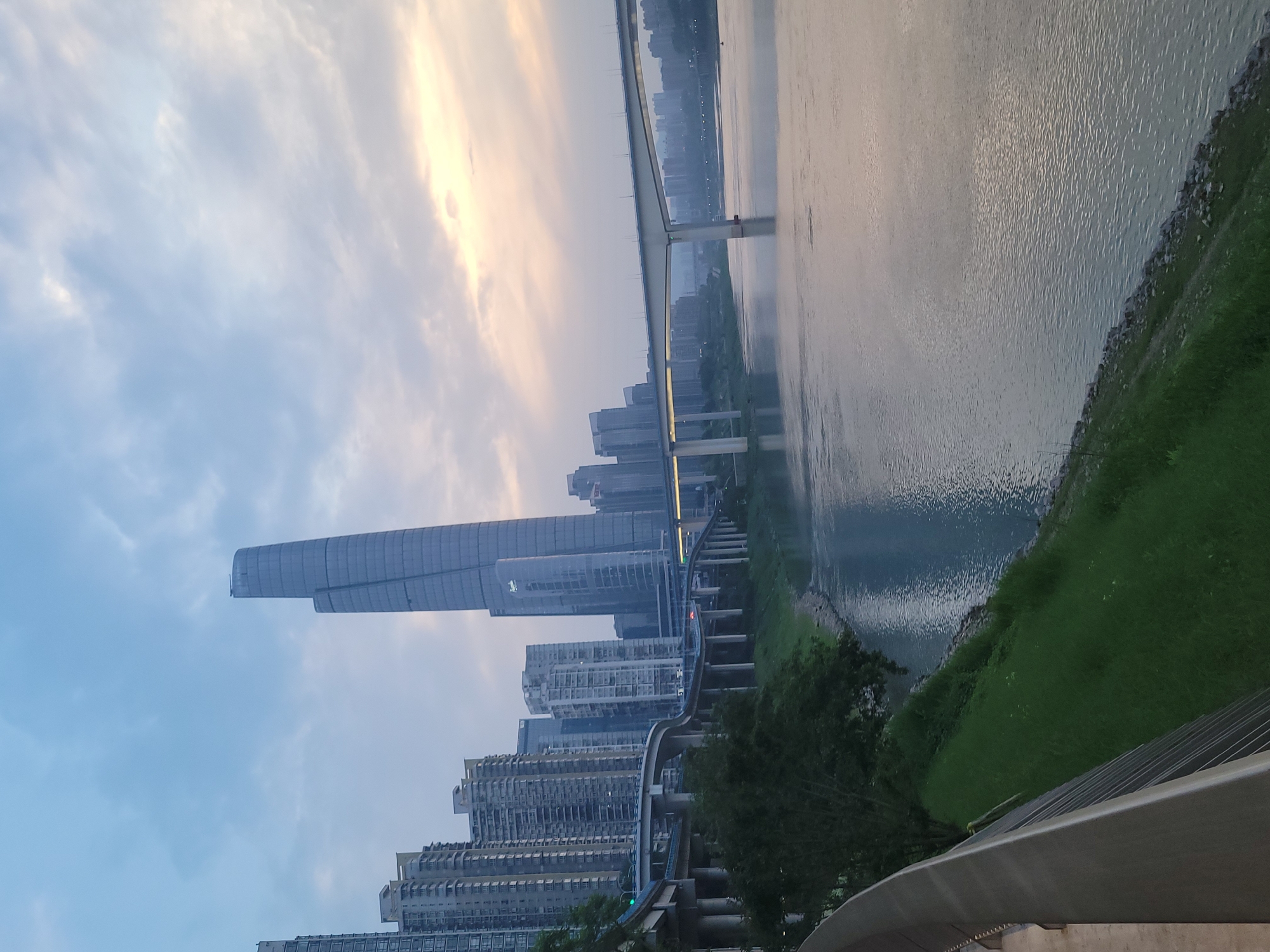 The image depicts a cityscape along a riverbank, likely in Yuzhong District, China, evidenced by the presence of modern high-rise buildings, a distinctive skyscraper, and a bridge. A bridge spans over the river. The sky is overcast, with the sun's rays visible behind the clouds. Green vegetation grows along the riverbank, and an elevated structure, possibly a railway or walkway, runs along the buildings.