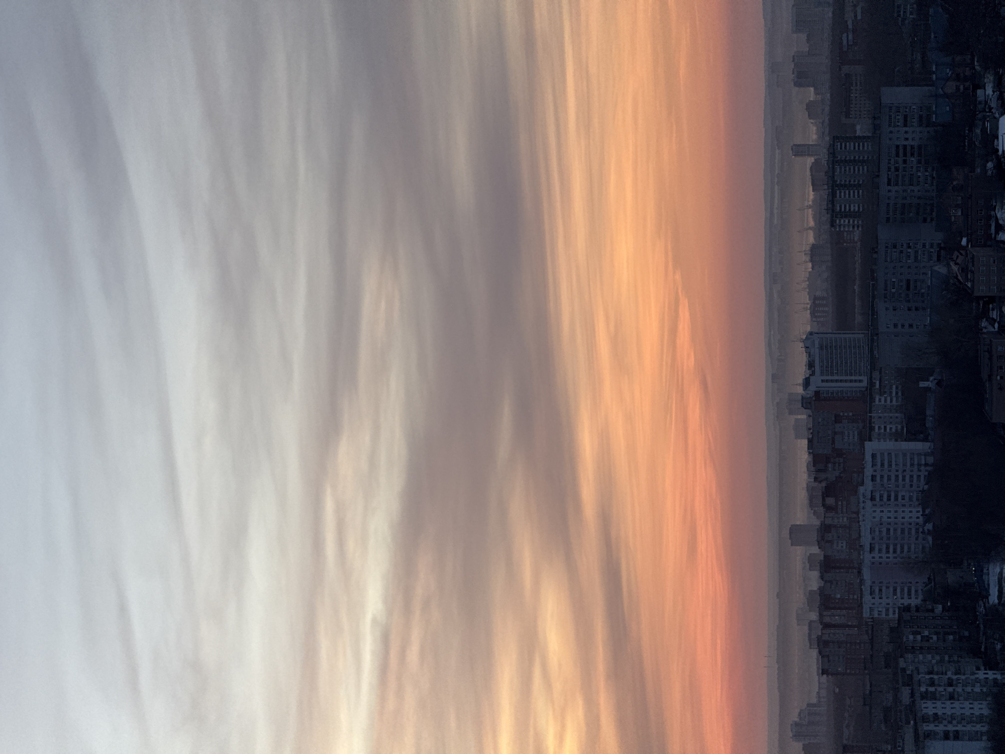 The image displays an expansive high-angle view of the city of Perm, Russia, at either sunrise or sunset. The sky occupies approximately the upper two-thirds of the frame, exhibiting a prominent color gradient. It transitions from a pale grey-blue at the top to a muted yellow-orange, then deepening to a vivid reddish-orange directly above the horizon line. Wispy, elongated clouds are visible across this gradient.
Below the sky, the cityscape extends into the distance, partially obscured by atmospheric haze. The foreground and midground are dominated by numerous multi-story residential apartment buildings, rendered primarily as dark silhouettes against the lighter sky. These structures appear to be made of concrete or similar materials, displaying repetitive window patterns. Interspersed among the buildings are dark patches of what appears to be bare deciduous vegetation.
In the far distance, beyond the immediate urban density, additional high-rise buildings and what appear to be industrial structures, possibly cranes, are faintly discernible through the haze, suggesting proximity to a major body of water, likely the Kama River. No individuals, discernible actions, or interactions are visible within the frame. The overall scene presents an urban environment under diffuse, colorful twilight illumination.