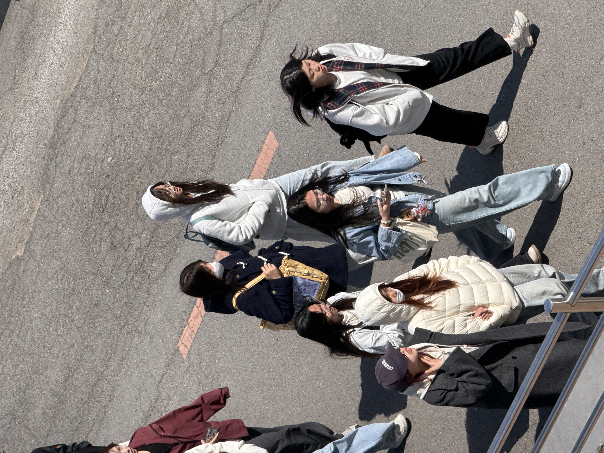 An overhead shot shows six people lying on an asphalt surface. A painted peach-colored line is visible near the center of the image. The people appear to be grouped casually, with varied clothing and postures. An object possibly a metal rail of some kind is in the lower right. The location is the city of Zhongtun, China.