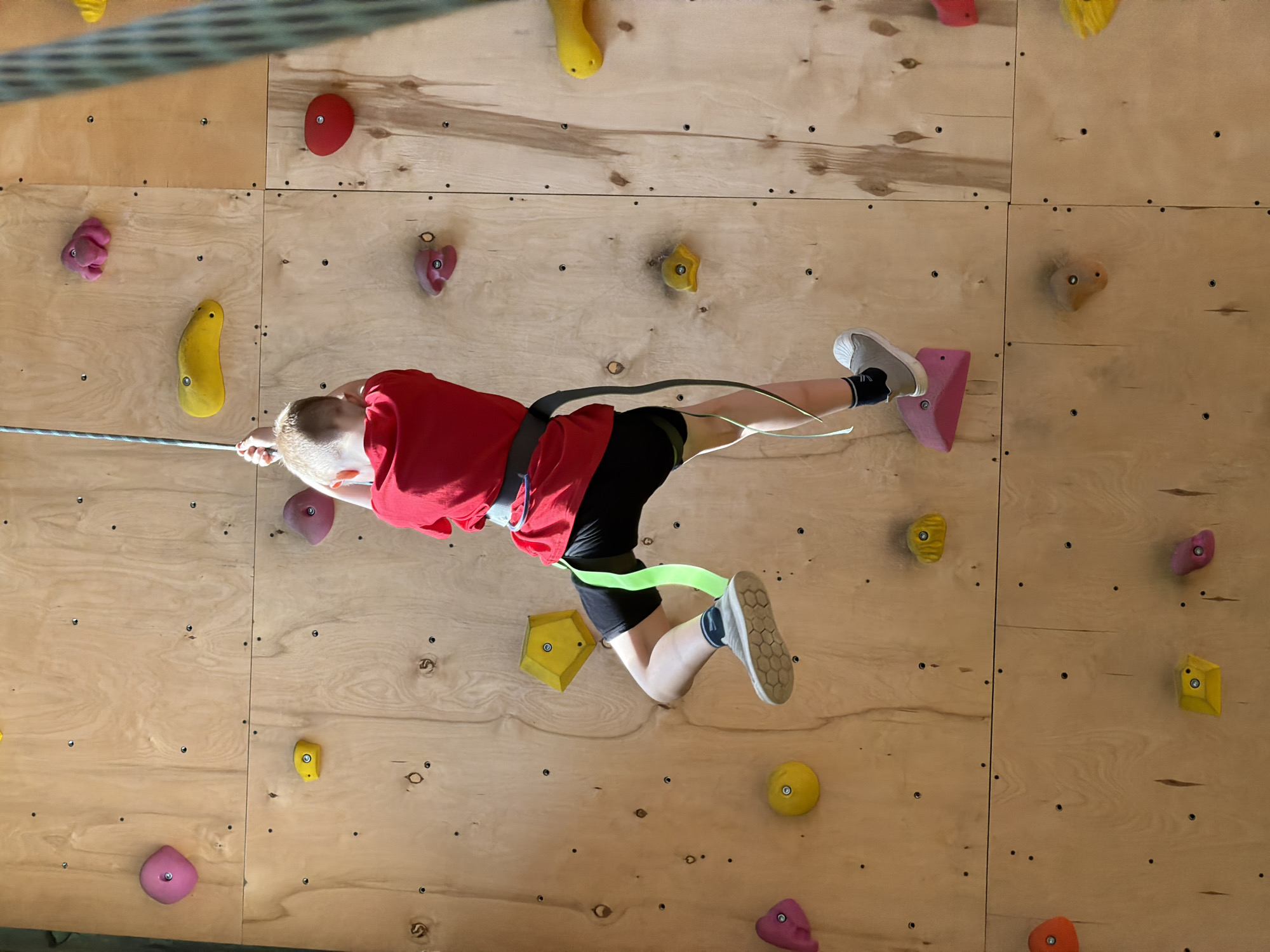 A boy is rock climbing indoors on a wooden climbing wall. The wall has various colored climbing holds attached to it. The boy is wearing a red t-shirt, black shorts, and gray shoes. He is wearing a climbing harness and is attached to a rope. The background is the wooden climbing wall. The location is unknown, and there is no information to suggest it is in Izhevsk, Russia.