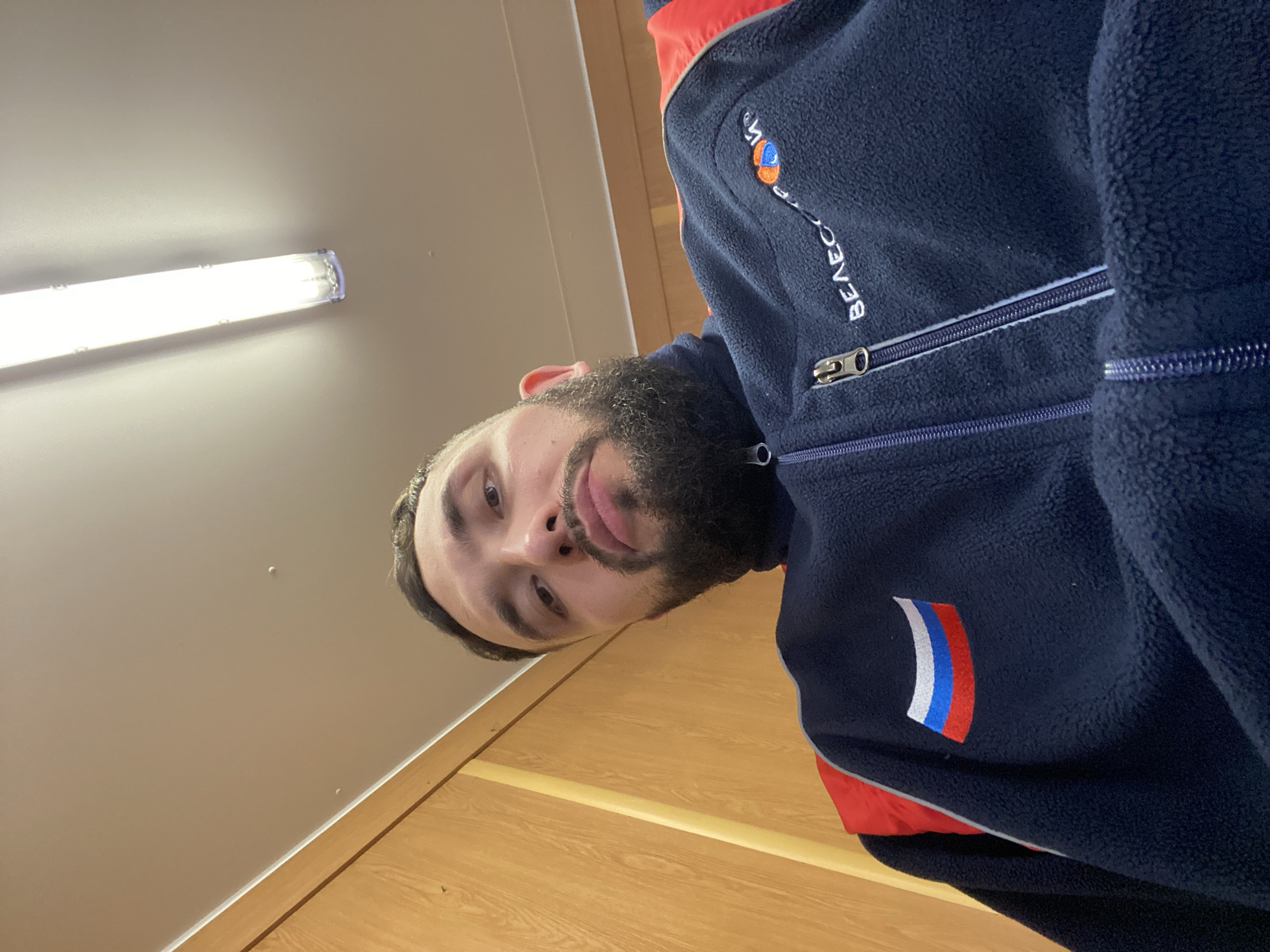 A man with a dark beard and short dark hair is seen from a slightly low angle, wearing a dark blue fleece jacket with red accents. The jacket has a Russian flag emblem on the left breast and the word "BEAECC" vertically embroidered on the left upper chest, along with a logo above the word. He is looking directly at the camera with a neutral expression.
The background consists of a light-colored wall with a fluorescent light fixture at the top left and light wooden paneling at the bottom.
It is not possible to identify the location as the city of Luzhitsy, Russia based on the available information in the image.