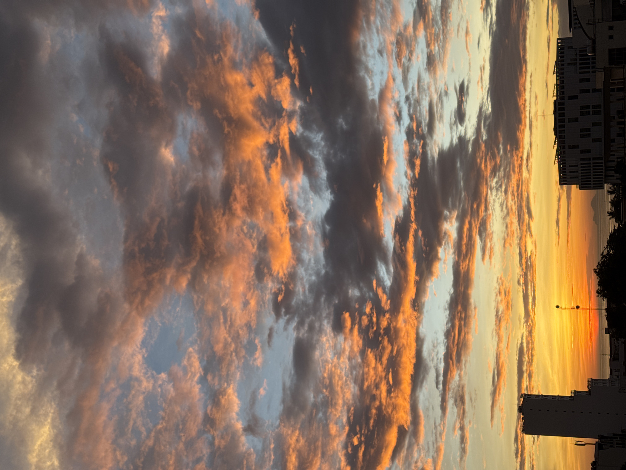 A dramatic sunset paints the sky over Nha Trang, Vietnam, with vibrant hues of orange, pink, and grey. Dark, brooding clouds are interspersed with streaks of fiery orange, while patches of soft blue peek through. In the foreground, the silhouettes of buildings are visible against the radiant sky. To the left, a tall, rectangular building stands prominently, its structure stark and defined. To the right, a more modern, flat-topped building with numerous windows is partially illuminated by the sunset. A transmission tower with multiple antennas rises from the cityscape, its metallic structure adding a vertical element. Distant islands are faintly visible on the horizon, merging with the sea under the colorful twilight. The overall impression is one of natural beauty and urban development coexisting.
