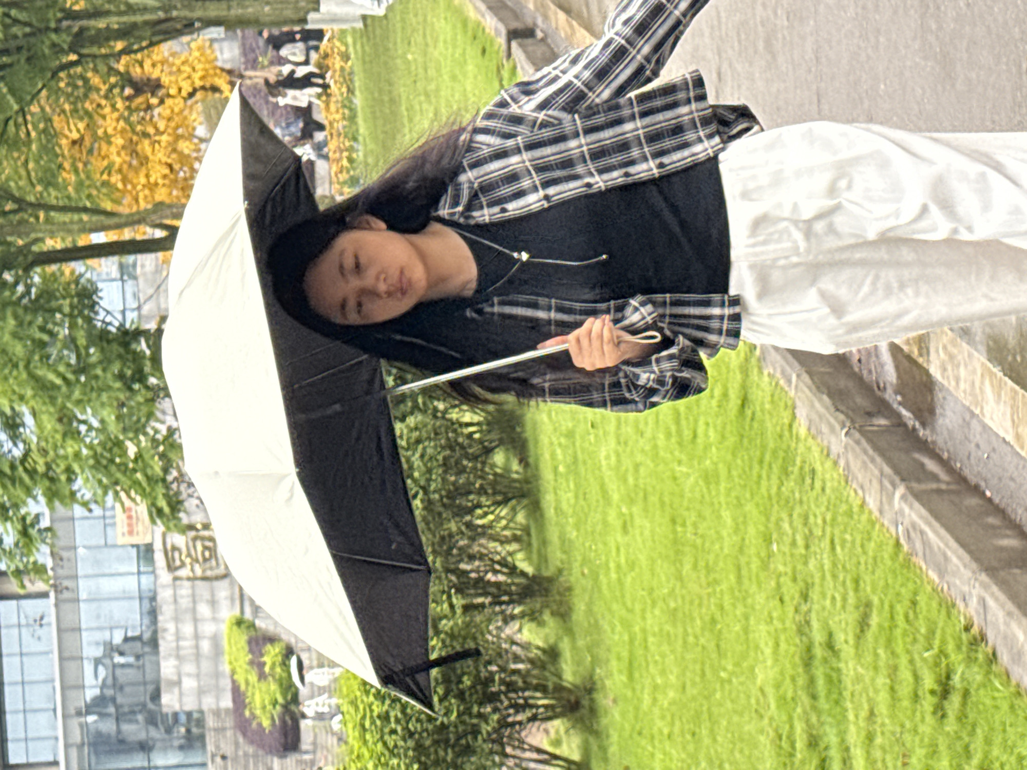 A young woman is walking on a sidewalk with an umbrella. She is wearing white pants, a black shirt, and a black and white plaid shirt. The sidewalk is next to a patch of green grass and a curb. In the background, there are trees, grass, a building with signage in Chinese characters, and people walking in the distance. Based on the signage, the location is likely Jiangjin, China.