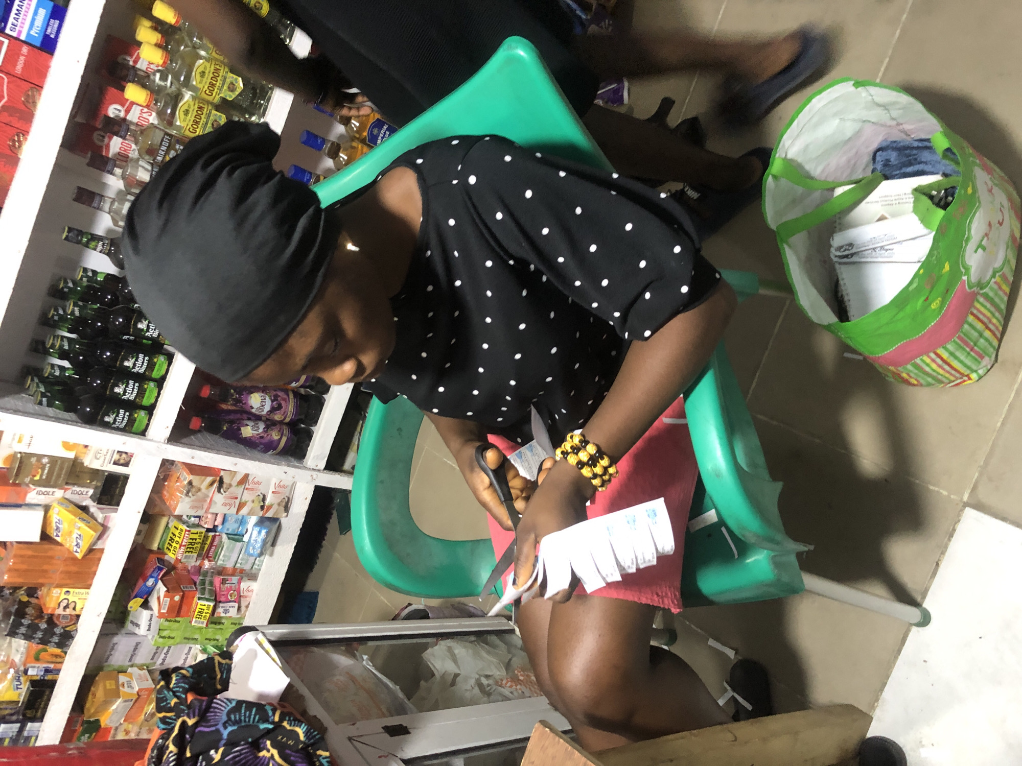 In Ado, Nigeria, a young woman sits on a green plastic chair, wearing a black headscarf and a black polka-dot top. She is actively cutting strips of white paper with black-handled scissors, holding the paper in her left hand and the scissors in her right. Her right wrist is adorned with a beaded bracelet. A pink cloth is draped over her lap. To her left, a wooden table is visible, and a colorful bag filled with items sits on the floor. The background reveals shelves stocked with various products, including bottled beverages and boxed goods, suggesting a retail environment.