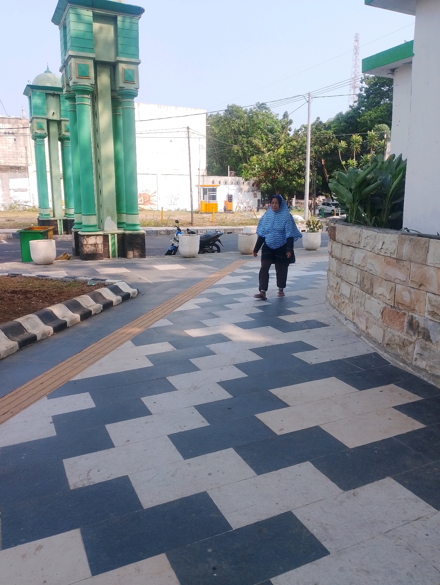 A woman wearing a blue and white patterned headscarf and a dark blue top walks on a pedestrian walkway in Probolinggo, Indonesia. The walkway is paved with large rectangular tiles arranged in a geometric pattern of alternating dark grey and light beige. A textured yellow tactile paving strip runs along the left edge of the walkway. To the left of the walkway is a low, black and white striped curb bordering a patch of dry brown earth. Beyond that, a street is visible with a green trash bin and a white planter. In the background, there are several tall, green, pillar-like structures with domed tops. Across the street, a white building with some graffiti and a small yellow structure are visible, along with trees and a cell tower in the distance. The woman is walking away from the camera, and there are no visible interactions between her and any other individuals. The sky is clear and blue.