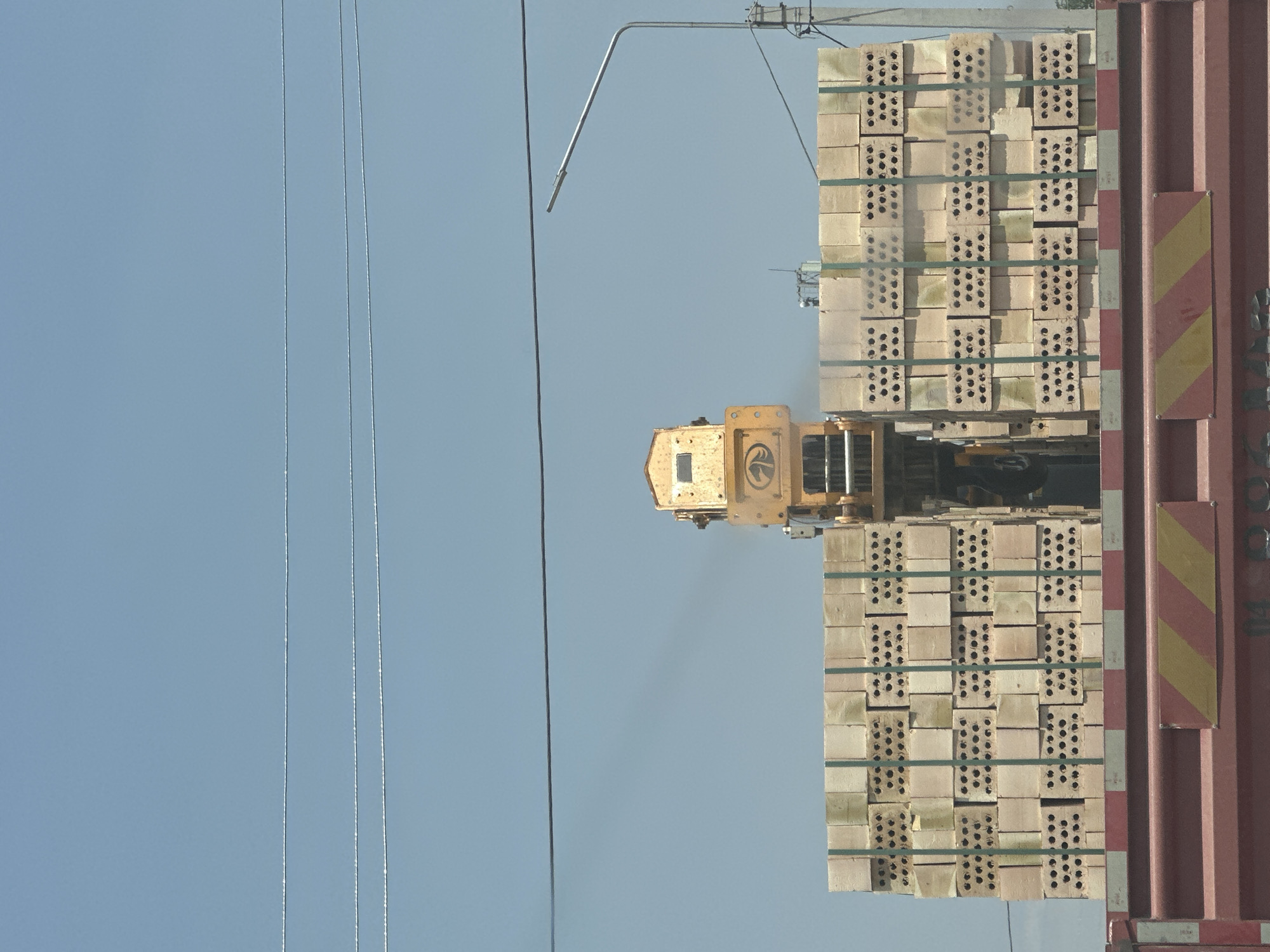 This media file shows a forklift operating near a stack of bricks. The forklift, which is yellow, is positioned in the center of the frame. A large stack of light-colored bricks, bound with green strapping, is to the right of the forklift. The bricks are neatly arranged, with each brick exhibiting multiple circular holes. To the far right of the image is the side of a large, reddish-brown container. There are two hazard stickers on the container: one with diagonal red and white stripes, and another with diagonal yellow and red stripes. The background is a clear, pale blue sky. Three thin, dark cables run vertically from the top to the bottom of the left side of the frame, suggesting the presence of power lines or similar infrastructure. The lighting suggests it is daytime. The scene appears to be an outdoor industrial or logistics area, possibly a port or a construction site. There is no visible text in the image. The orientation of the image is landscape.