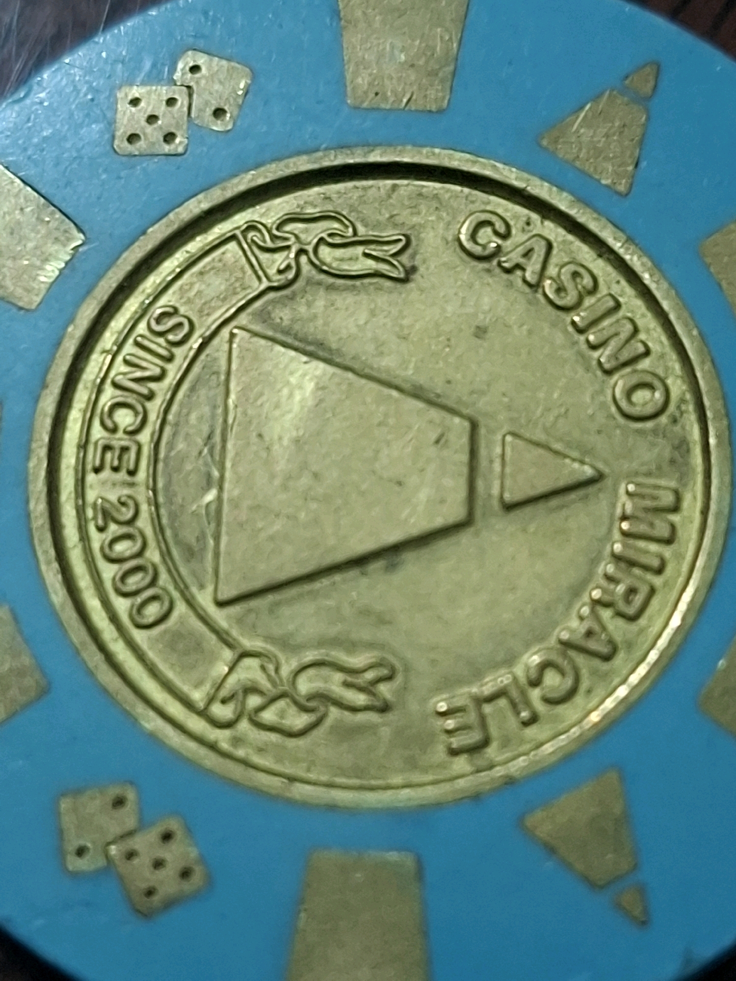 The image is a close-up of a casino chip. The chip is turquoise with gold accents. In the center of the chip is a gold emblem that appears to be a stylized play button or arrow. Encircling the emblem is text that reads "SINCE 2000" on the left side and "CASINO MIRACLE" on the right side, both in gold lettering. There are also small gold dice symbols on the left side of the chip, near the top and bottom edges. The background is the turquoise surface of the chip itself, with some gold trapezoidal shapes visible around the edge. There is no information in the image to suggest its location in Almaty, Kazakhstan.