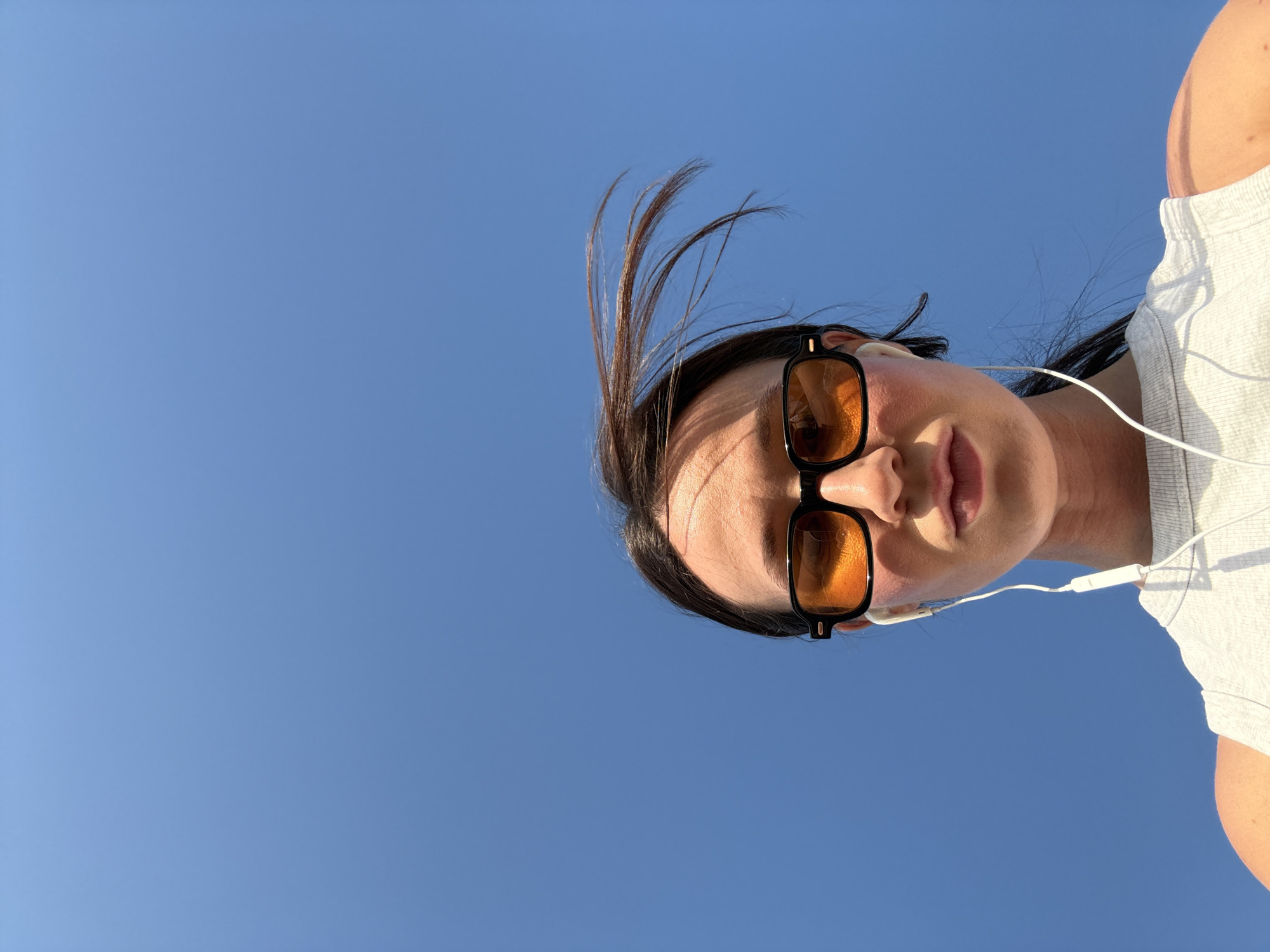 An outdoor, low-angle portrait captures a person's upper body against a clear, expansive blue sky. The image is oriented with the person's head and shoulders positioned in the upper right, looking down towards the camera, which suggests a selfie taken from a worm's-eye view. The subject wears black-framed sunglasses with warm, orange-tinted lenses that reflect bright sunlight. White wired earphones are in use, with one earpiece visible in the left ear and the wire extending downwards. Dark hair, possibly in a ponytail or bun, has several strands visibly blown upwards by the wind, creating a dynamic effect against the serene sky. The person is dressed in a light-colored, possibly ribbed, short-sleeved top. The bright, uniform blue background indicates a sunny day with clear weather. No text is visible in the image. The scene is located in Tel-Aviv, Israel, as per context.