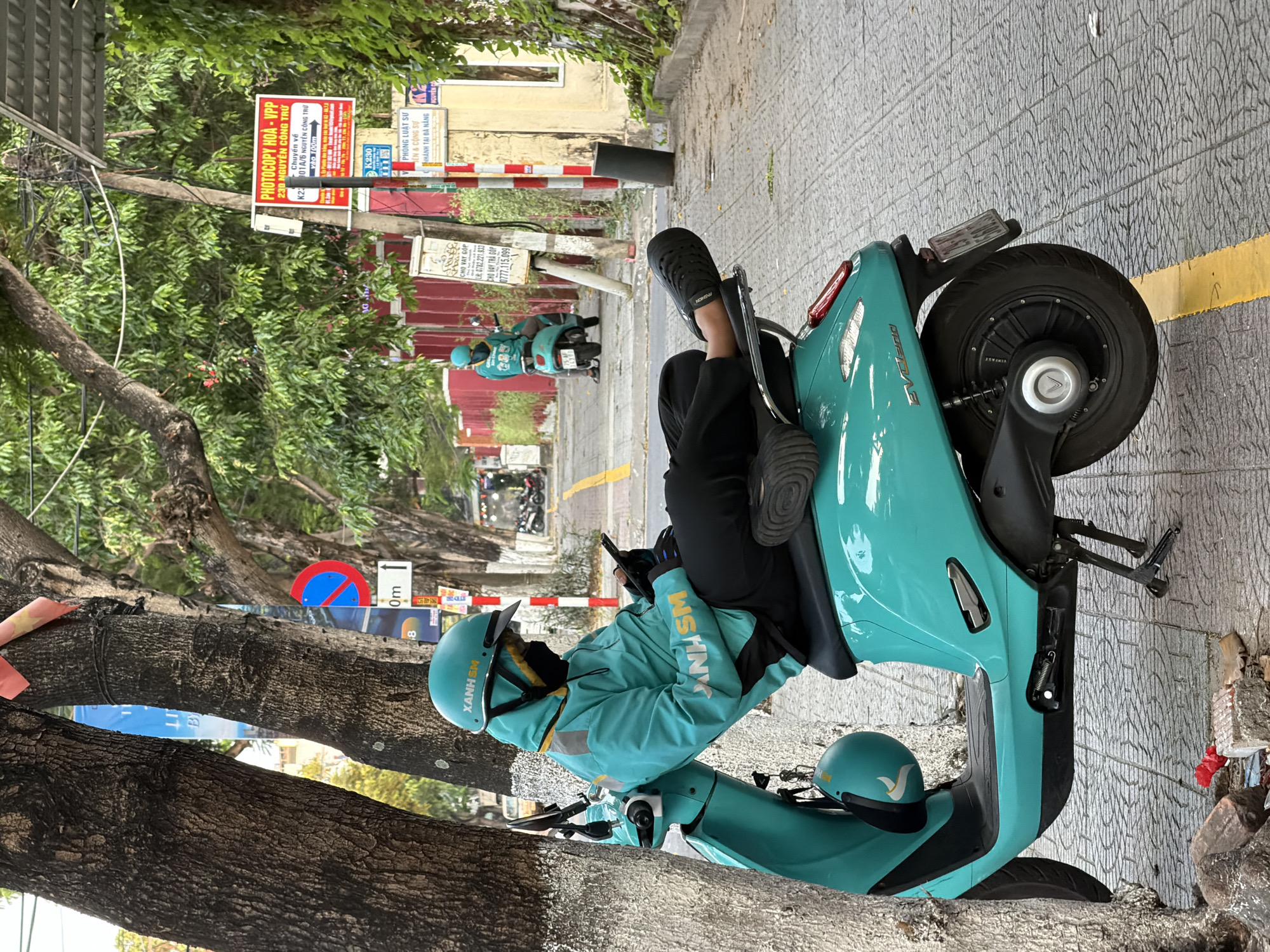 The image displays a daytime street scene in Đà Nẵng, Vietnam, centered around a person in a distinctive teal uniform associated with the "XANH SM" electric ride-hailing and delivery service. The individual, wearing a matching jacket and helmet, sits relaxed on a teal electric scooter, with their feet propped up, seemingly engaged with a mobile phone. Another identical "XANH SM" helmet rests on the ground beside the scooter.
The setting is an urban street lined with mature trees providing a leafy canopy. In the background, further down the road, another "XANH SM" driver on a similar scooter is visible. The street features a paved sidewalk and buildings.
Prominent visible text on street signs and banners includes "PHOTOCOPY HOÀ - VPP" (Photocopy Hoà - Stationery) with addresses like "230 NGUYỄN CÔNG TRỨ" and "101/6 NGUYỄN CÔNG TRỨ". Another sign advertises a "PHÒNG LUẬT SƯ" (Law Office) with a "NHÁNH TẠI ĐÀ NẴNG" (Branch in Da Nang). Signs offering "CHO VAY GÓP" (Installment Loans) with contact numbers are also present. A blue circular sign with a red diagonal line indicates a "no parking" or "no stopping" zone. The scooter has "EVOLUS" emblazoned on its side, likely its model name. The scene captures a moment of rest for a driver in a modern Vietnamese city environment, characterized by active electric transport services.