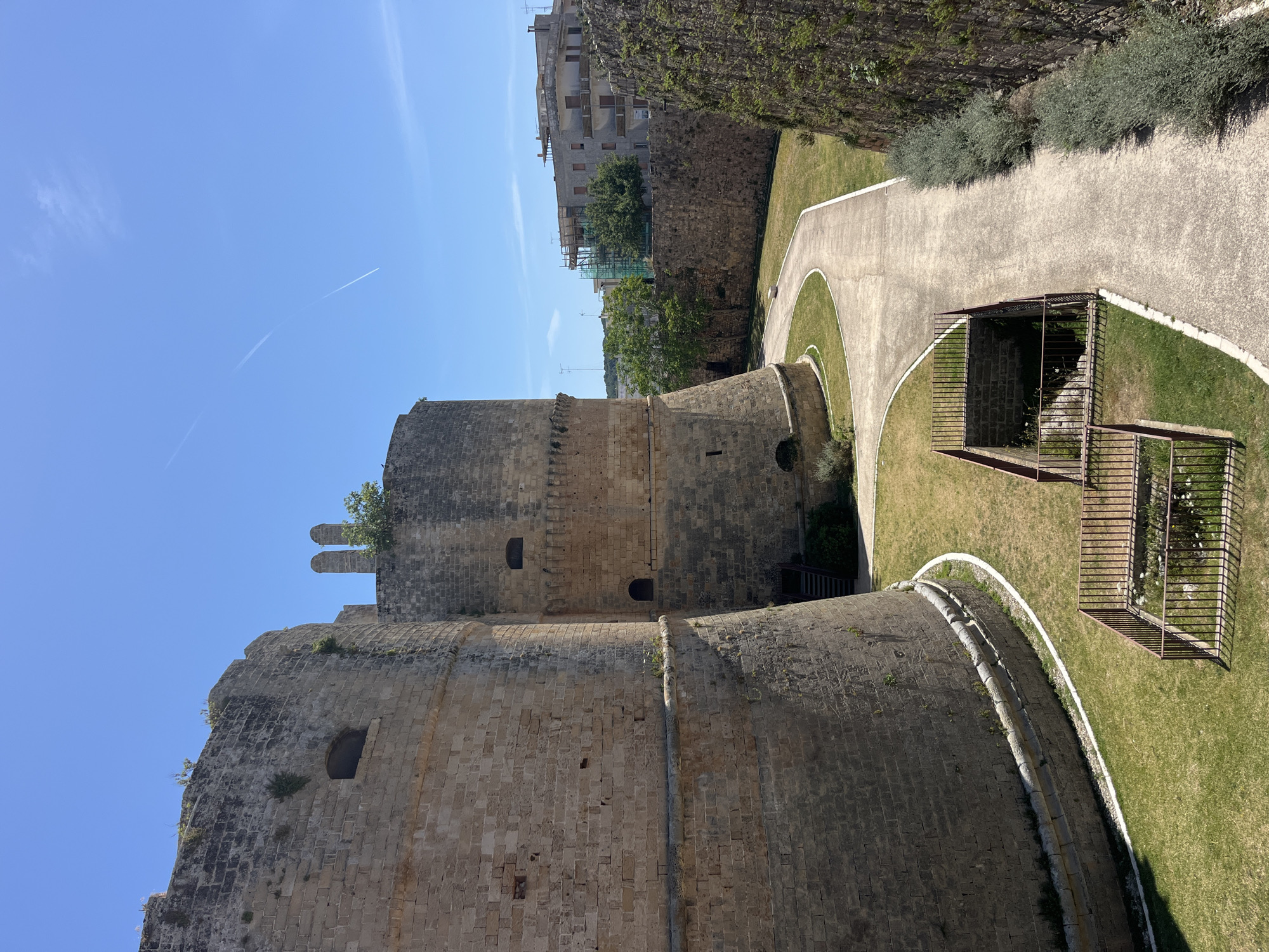 This image shows the Aragonese Castle in Otranto, Italy. The castle is made of light-colored stone and has a cylindrical tower. A paved walkway curves around the castle. There is a grassy area with a metal cage-like structure. The sky is clear and blue. Other buildings and trees are visible in the background.