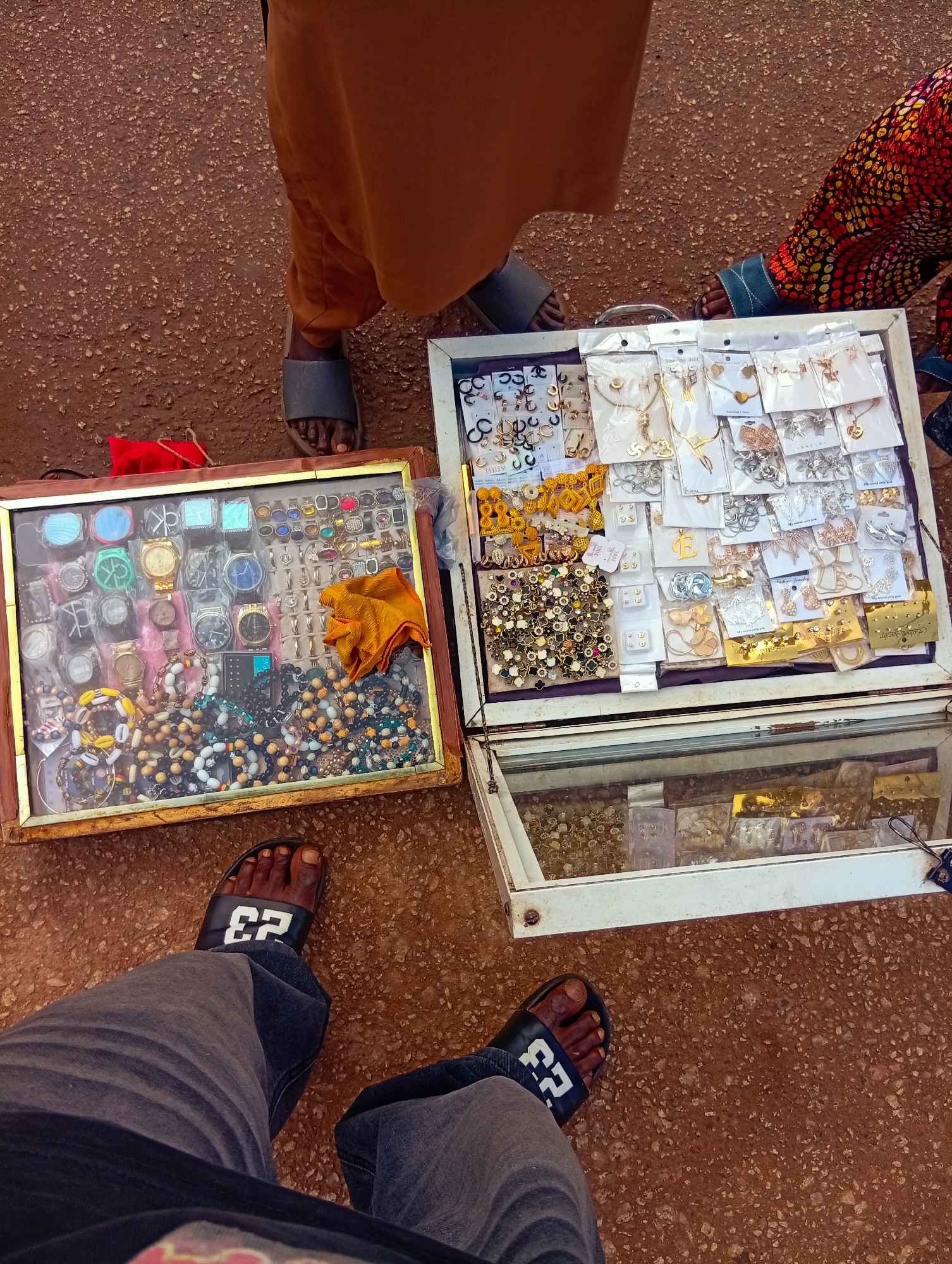 In Bukuru, Nigeria, a street vendor displays an array of jewelry and watches for sale. Two display cases are visible, one rectangular with a wooden frame and the other a white display case with a glass top.
The wooden-framed case is filled with an assortment of watches, each individually packaged or displayed. Below the watches, a variety of beaded necklaces and bracelets are arranged, some featuring colorful beads and cowrie shells. An orange cloth lies on top of some of the jewelry.
The white display case showcases a collection of packaged jewelry, including necklaces, earrings, and pendants. These items are neatly arranged in plastic packaging, with some displaying price tags or brand names. The lower section of this case is also filled with more jewelry items in plastic packaging.
Several individuals are present, their legs and feet visible in the foreground and around the displays, suggesting they are either the vendor or potential customers. One person wears dark gray jeans and black slides with "23" printed in white. Another person's legs are visible in a brown dress or skirt and dark gray slides. A third person, wearing a colorful patterned garment, also has their legs and feet visible.
The ground beneath the displays appears to be a reddish-brown, rough surface, possibly a dirt road or unpaved area. The overall scene suggests an outdoor market or roadside stall, likely during daylight hours. The atmosphere appears to be one of commerce, with the focus on the goods being sold.
Visible text includes the number "23" on the footwear and some labels on the jewelry packaging, though the specific content of these labels is not legible.