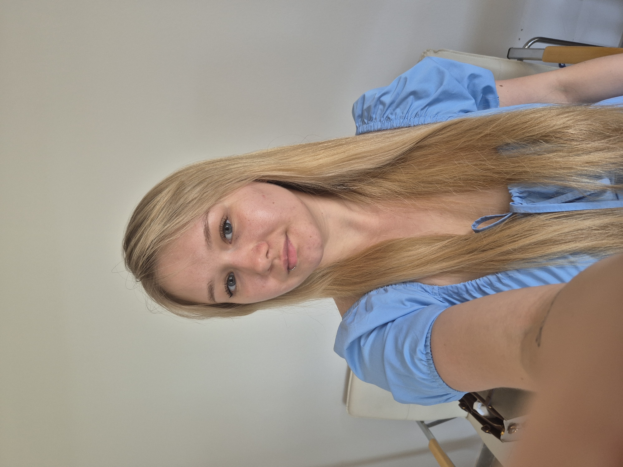 A young blonde woman with blue eyes and a piercing on her lower lip is sitting in a chair. She is wearing a blue puff-sleeved dress. The background is a plain white wall. There is a chair and a table in the background. The image appears to have been taken indoors. There is no indication of this location being in Chaikovsky, Russia.