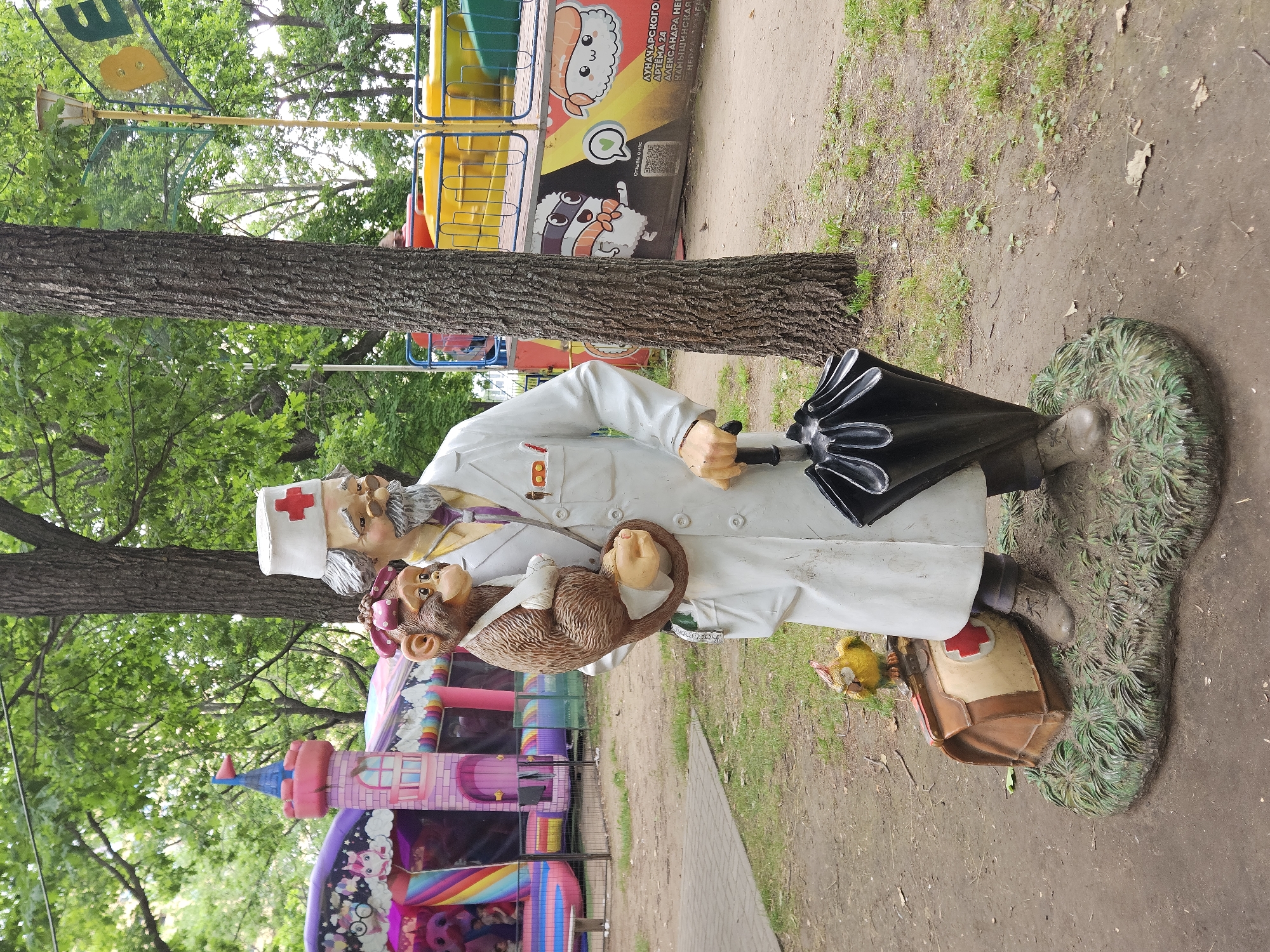 A statue of a doctor holding a monkey stands in a park in Ulyanovsk, Russia. The doctor is wearing a white lab coat and a cap with a red cross. The monkey is wearing a bandage on its arm. A medical bag and a small bird statue are at the doctor's feet. In the background, a bounce house and amusement park rides are visible.
