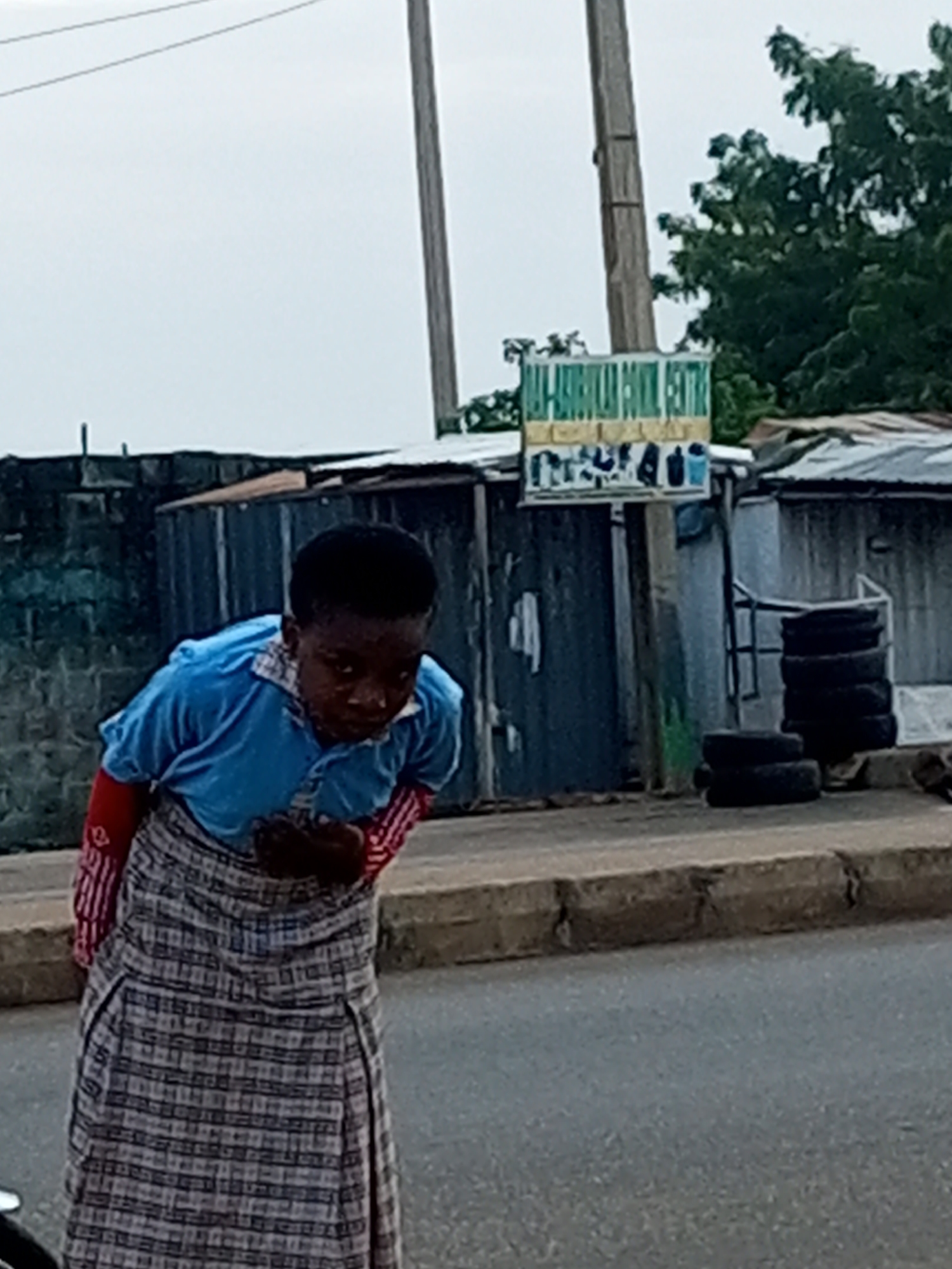 The image displays a street scene under an overcast sky. A single individual, appearing to be a young person or child with dark skin, stands centrally. They are dressed in a light blue short-sleeved polo shirt worn over a red long-sleeved undershirt, and a long, plaid skirt in grey and white. The individual is bent forward at the waist, looking downwards, with their hands held in front of their abdomen.
The foreground consists of an asphalt road, bordered by a concrete curb on the right. The background features various urban structures. To the left, a dark, possibly concrete block wall is visible. In the mid-ground, a tall wooden utility pole supports a rectangular sign. The sign's upper section is green with partial white text, including "CENTRE" at the end, while the lower section displays images of electronic devices and other items. Behind the pole and extending to the right are several structures built from corrugated metal sheeting, some appearing weathered. A large, leafy green tree is present in the upper right background, behind these structures. On the right side of the scene, two stacks of dark vehicle tires are visible, one tall and one shorter, positioned near the corrugated metal buildings. Two thin utility lines stretch across the upper left portion of the sky. The location is an unspecified city and country.