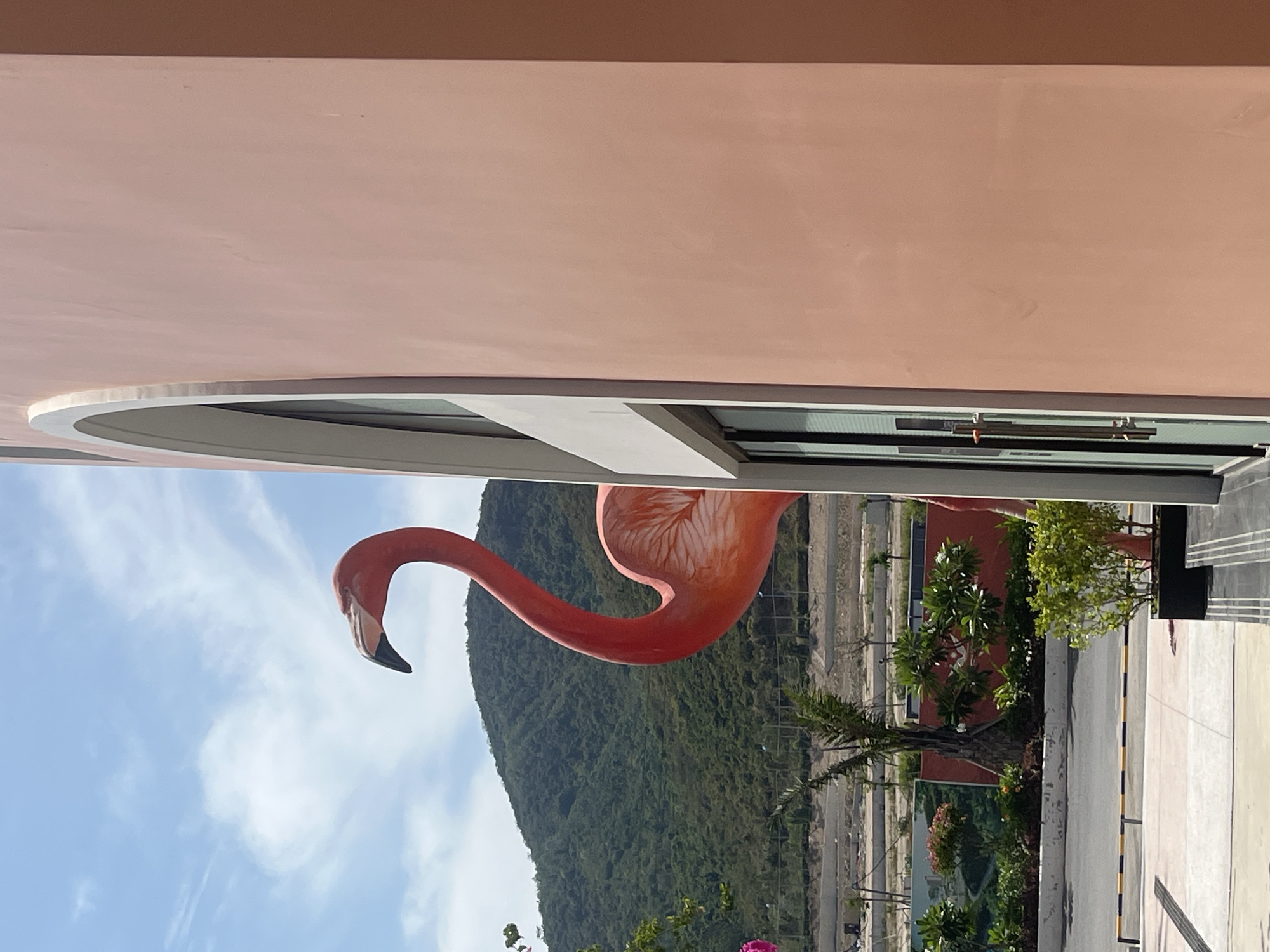 The photograph shows a large orange flamingo statue outside a building in Bắc Nha Trang, Vietnam. The statue is positioned in front of a green mountain range and a clear blue sky with some clouds. A building is visible in the foreground with a pink wall and a white curved architectural detail. Below the flamingo, there's a paved area with greenery, including trees and shrubs.