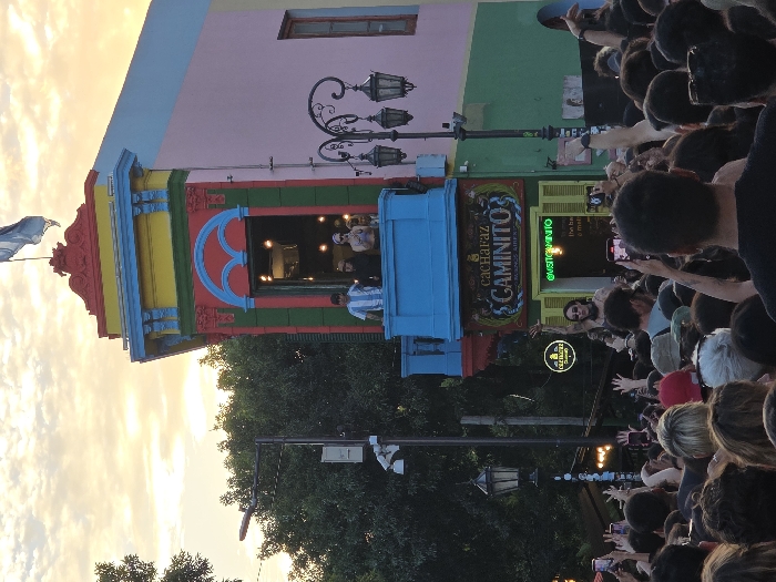 A vibrant, multi-colored building with intricate details is angled sharply against a sky with a golden hue, likely during sunset. In the building's doorway, people can be seen. Below, a crowd has gathered, capturing the scene with their phones. Streetlights and green trees are also present, completing the lively street view.