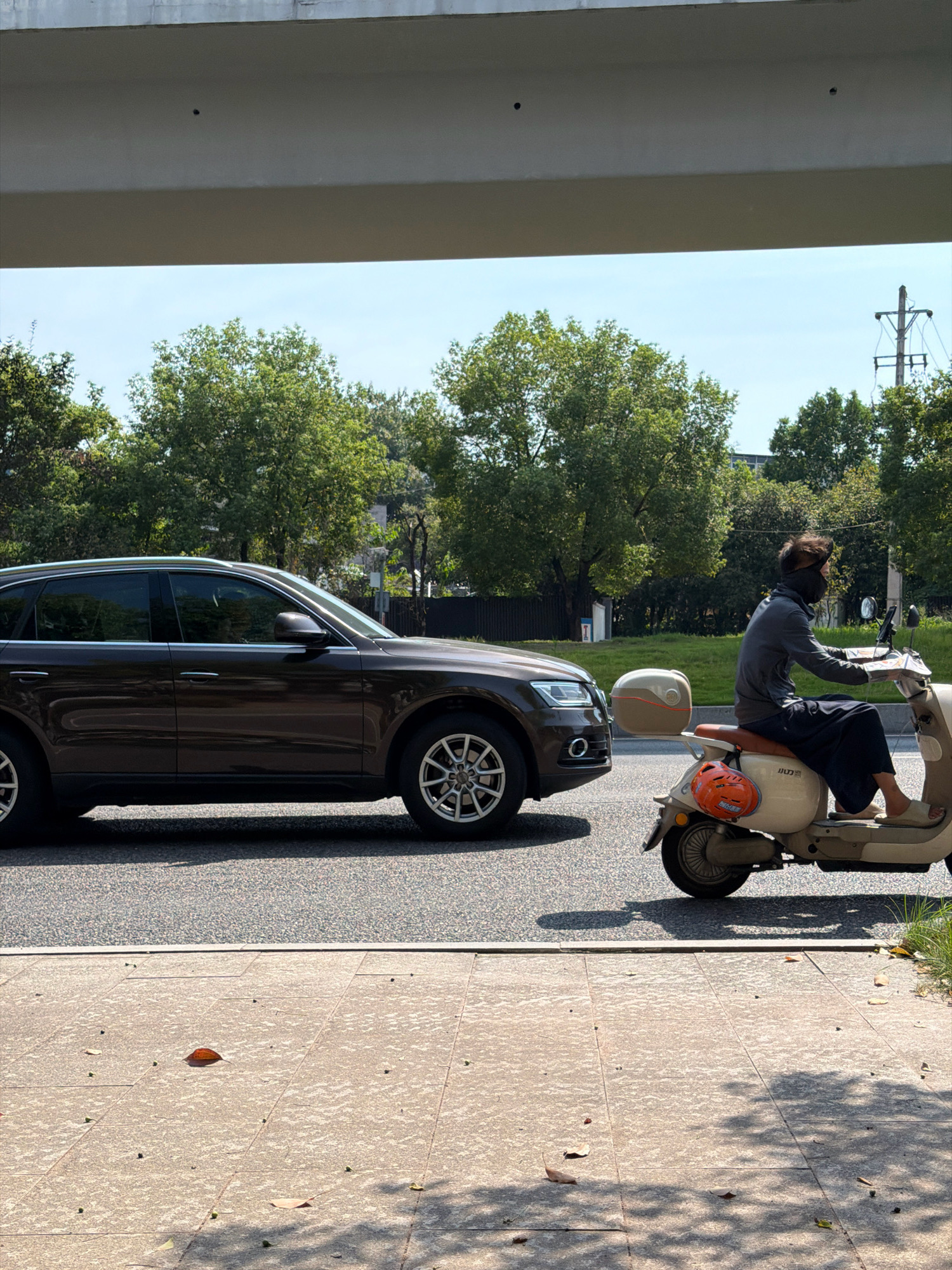 The image captures a street scene in Jiangjin, China, under a bright, clear sky. In the foreground, a paved sidewalk is visible, with scattered leaves and small dark objects. Beyond the sidewalk, a paved road runs horizontally. On the road, a dark brown SUV is parked, facing left. To the right of the SUV, a person wearing dark clothing and a black face mask rides a light-colored scooter. The scooter has an orange helmet attached to the back. Trees with dense green foliage line the background, with a concrete structure overhead suggesting an overpass or elevated roadway. The overall impression is a typical urban environment on a sunny day.