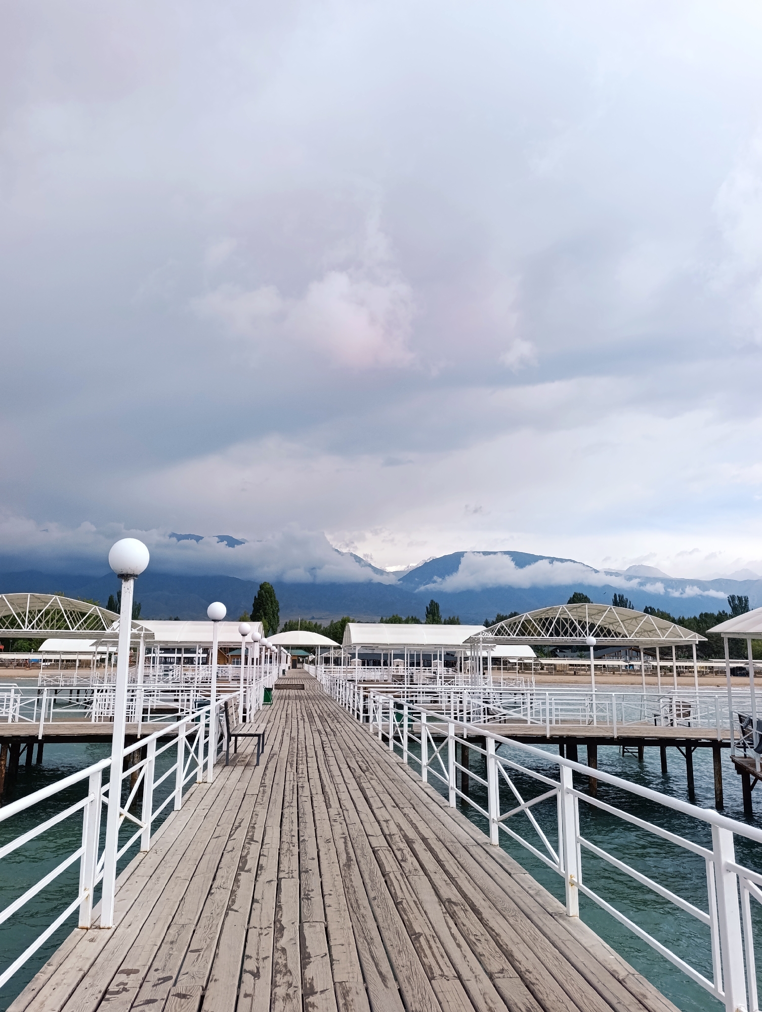 An outdoor, eye-level shot captures a wooden pier with white railings extending into a body of water. The pier is lined with white, open-air shelters. In the background, mountains are visible beneath a cloudy sky. The scene is located in the resort area of Sary-Oy, Kyrgyzstan.