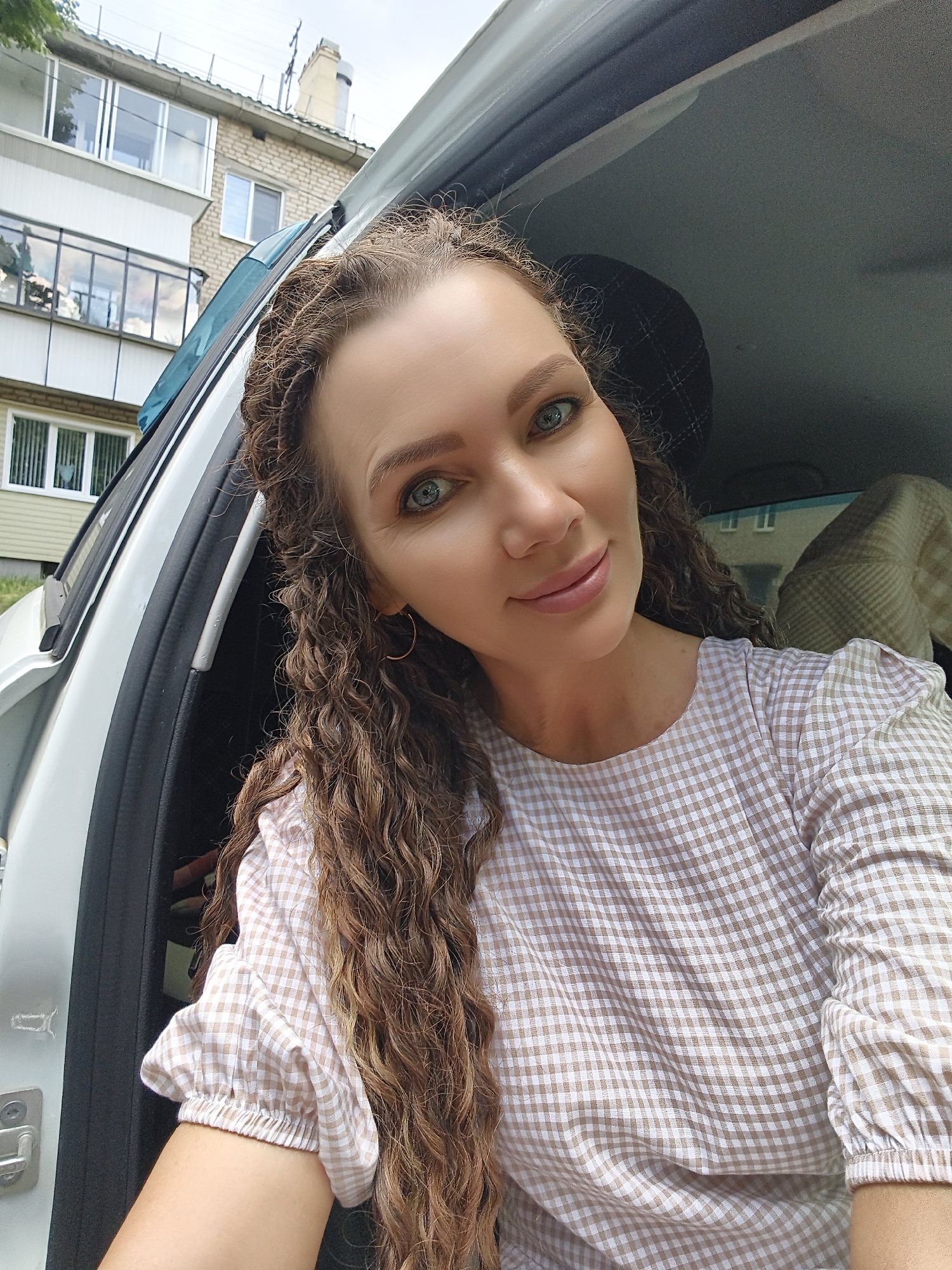 A medium shot captured from inside a vehicle shows a woman with long, curly brown hair and blue eyes. She is looking at the camera and smiling slightly. The woman is wearing a light-colored, checkered top. The vehicle's door is open, revealing a blurry background of a building and trees. The lighting suggests an outdoor, daytime setting.