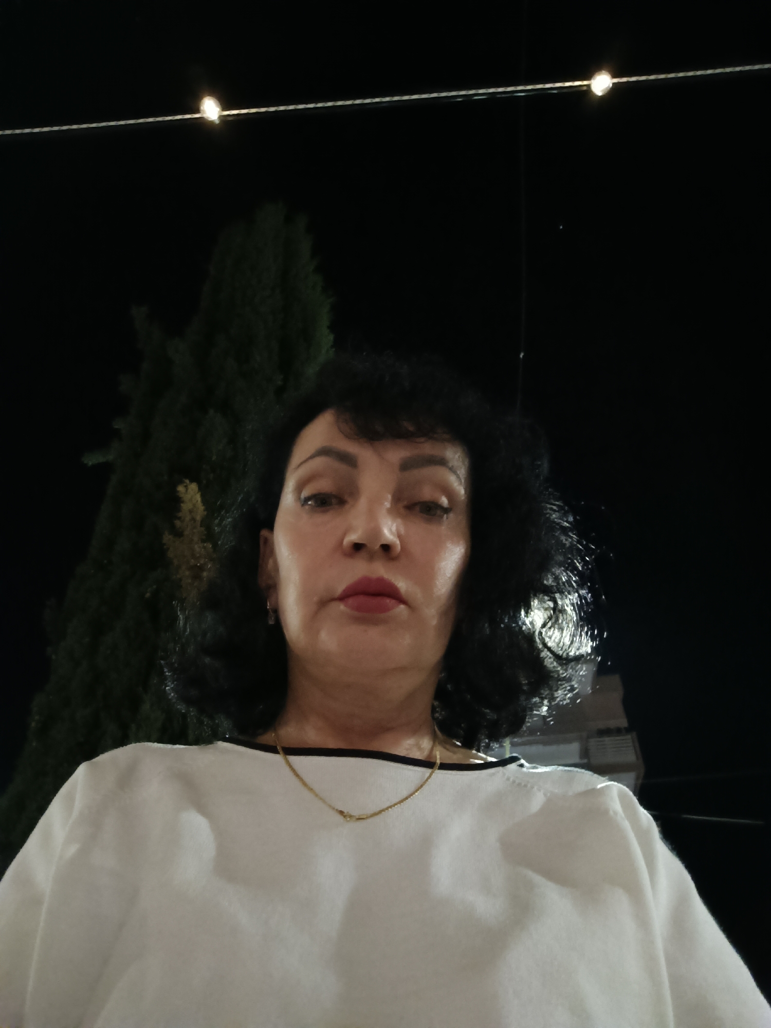 A medium shot shows a woman with dark, curly hair and red lipstick looking directly at the camera. She is wearing a white shirt and a gold necklace. The background is dark, with a string of lights visible above her and a dark green tree to her left. There is a building in the background to the right. The image appears to have been taken at night.