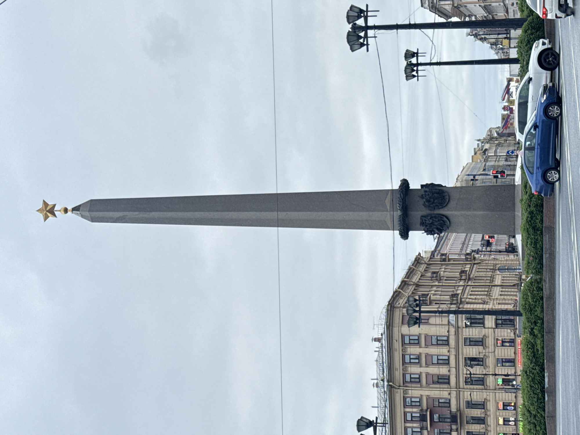 The image shows the Alexander Column in Palace Square in Saint Petersburg, Russia. The column is a tall, gray obelisk topped with a golden star. Buildings are visible in the background, as well as a street with parked cars. The sky is overcast. Street lights are also visible.