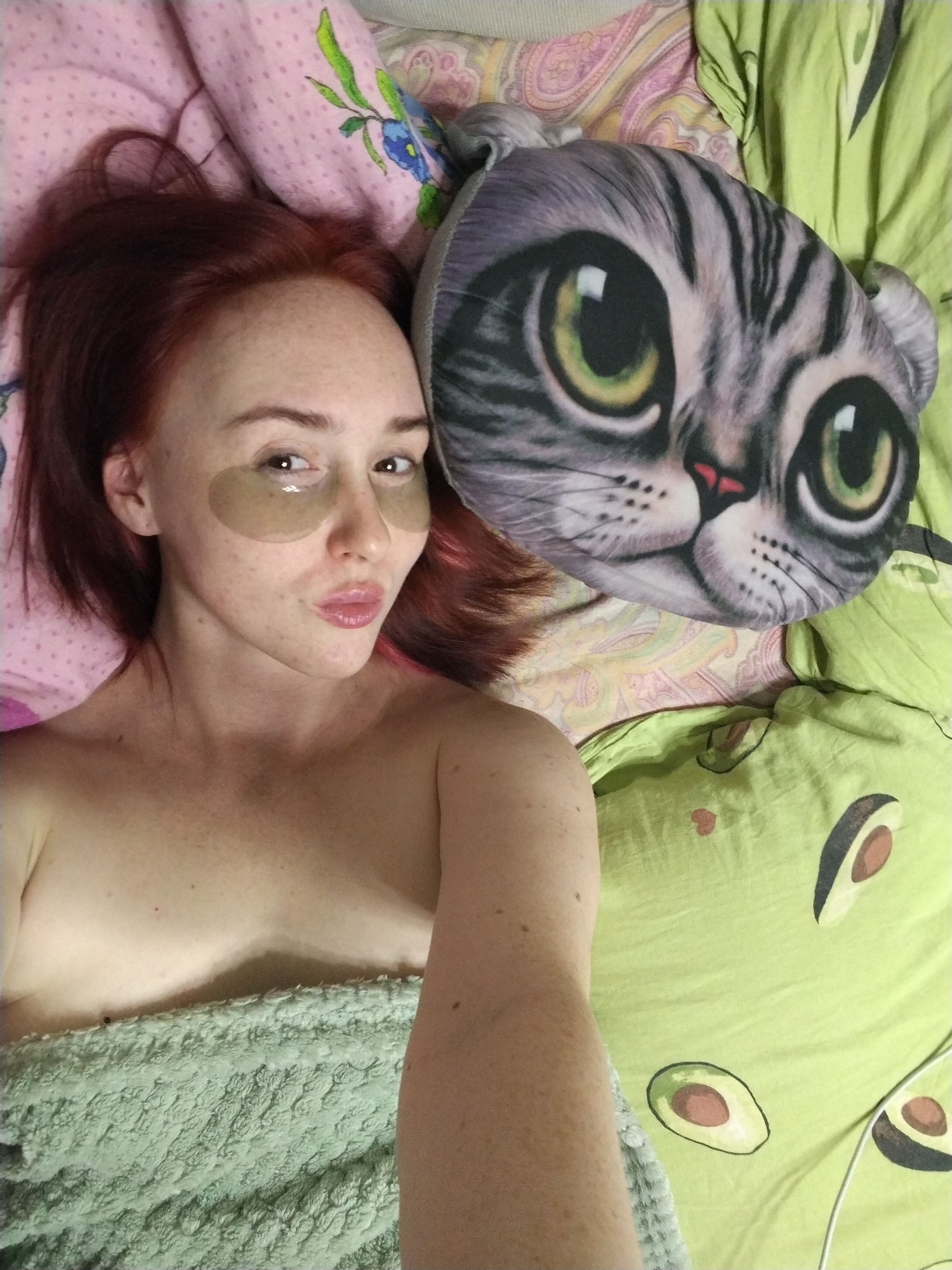 A red-haired woman with eye masks is lying in bed next to a cat-faced pillow. She is partially covered by a textured green blanket. The bed linens are a mix of patterns, including pink with floral designs, paisley, and light green with avocado prints. The location is not identifiable as Izhevsk, Russia from the visual information provided.