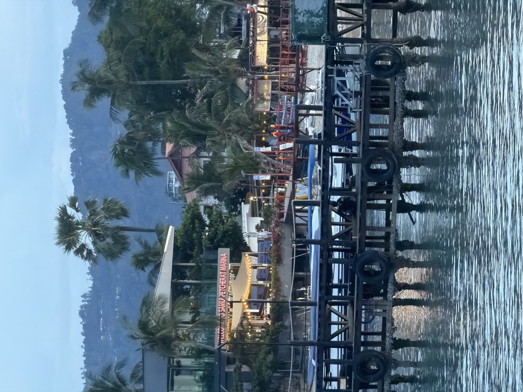 The image shows a waterfront scene at dusk in Baan Bang Rak, Thailand. In the foreground, a wooden pier extends over the water, supported by pilings encrusted with marine growth. Two tires are hanging from the pier's structure. Several boats are docked beneath the pier. In the background, a walkway with railings and lighting stretches along the shore, lined with palm trees and buildings. A sign reading "WELCOME TO SAMUI PETCHERAT MARINA" is illuminated with red lights. Further inland, lush green hills rise, dotted with lights from houses. The water is calm, reflecting the lights from the pier and surrounding structures. Two individuals wearing life vests are visible on the walkway, suggesting a marina or tourist area.