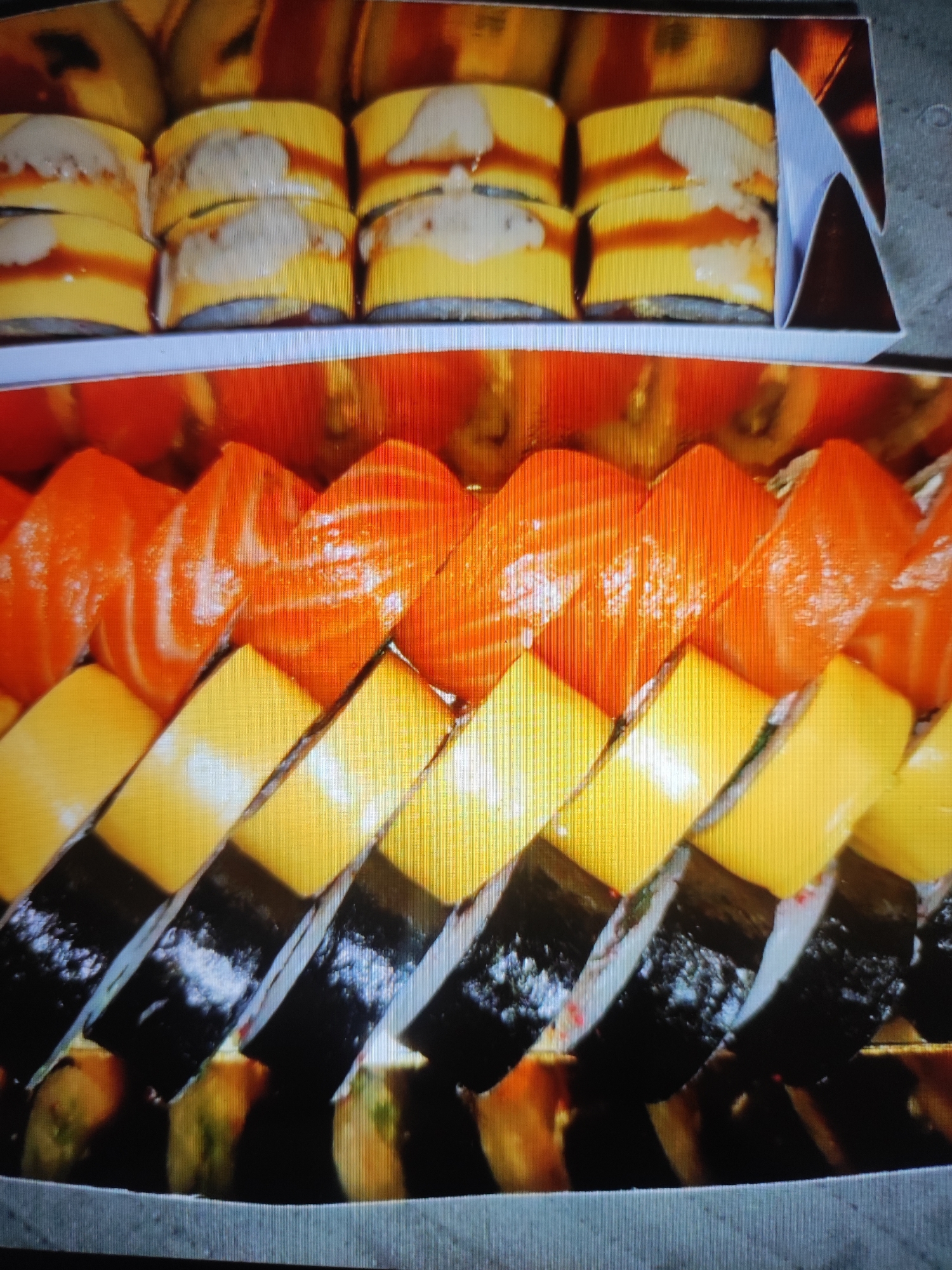 The image displays a close-up, overhead view of a sushi platter. The platter contains several rows of sushi rolls. The top row shows what appear to be egg rolls with a yellow outer layer, topped with a light-colored creamy sauce and a drizzle of dark sauce. Below that, a row of sushi rolls are visible with bright orange salmon on top, revealing the white flesh beneath. Beneath the salmon rolls are rows of sushi rolls with a distinct yellow layer, likely mango or avocado, paired with dark seaweed and white rice. The bottom-most visible layer shows the ends of the sushi rolls, revealing the rice and fillings, with some green specks. The sushi is arranged in a decorative, staggered pattern. The platter itself is set against a neutral, possibly grey, surface. There is no indication of the city of Cherkasy, Ukraine, in the image itself.