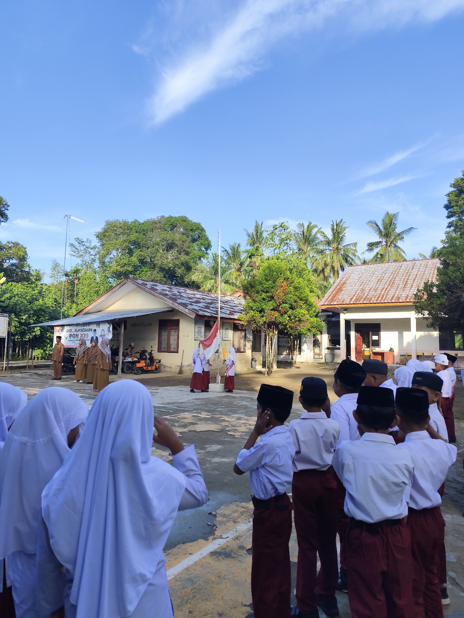 In the foreground, a group of young students, some wearing white hijabs and others wearing black caps, stand in an outdoor courtyard. They appear to be observing a flag-raising ceremony. In the middle ground, several students in uniform, some holding flags, are positioned near a flagpole. Behind them, a modest school building with a light-colored facade and a corrugated metal roof is visible. Palm trees and other lush greenery surround the school. The sky above is a clear blue with a few wispy clouds. The scene suggests a school event taking place on a bright, sunny day.
