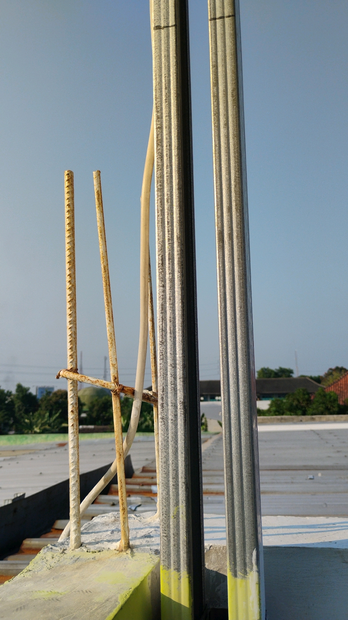 The image depicts a construction scene with a clear blue sky in the background. In the foreground, there are several vertical elements. Two prominent, ribbed metal poles stand side-by-side, with a white electrical conduit running between them. To the left of the metal poles are three rusty rebar rods, one of which is tied to the others with wire. These rebar rods are anchored in a concrete structure that is partially painted with bright yellow and lime green. In the mid-ground, a gray rooftop with some visible lines can be seen, with a dark angled structure extending upwards from the left. Beyond the rooftop, there is a line of green trees and some buildings with red tile roofs, suggesting a semi-urban or suburban environment. The overall scene is consistent with the ongoing construction of a building in Gumpang, Indonesia, under daylight conditions. There are no people or animals visible in the image.