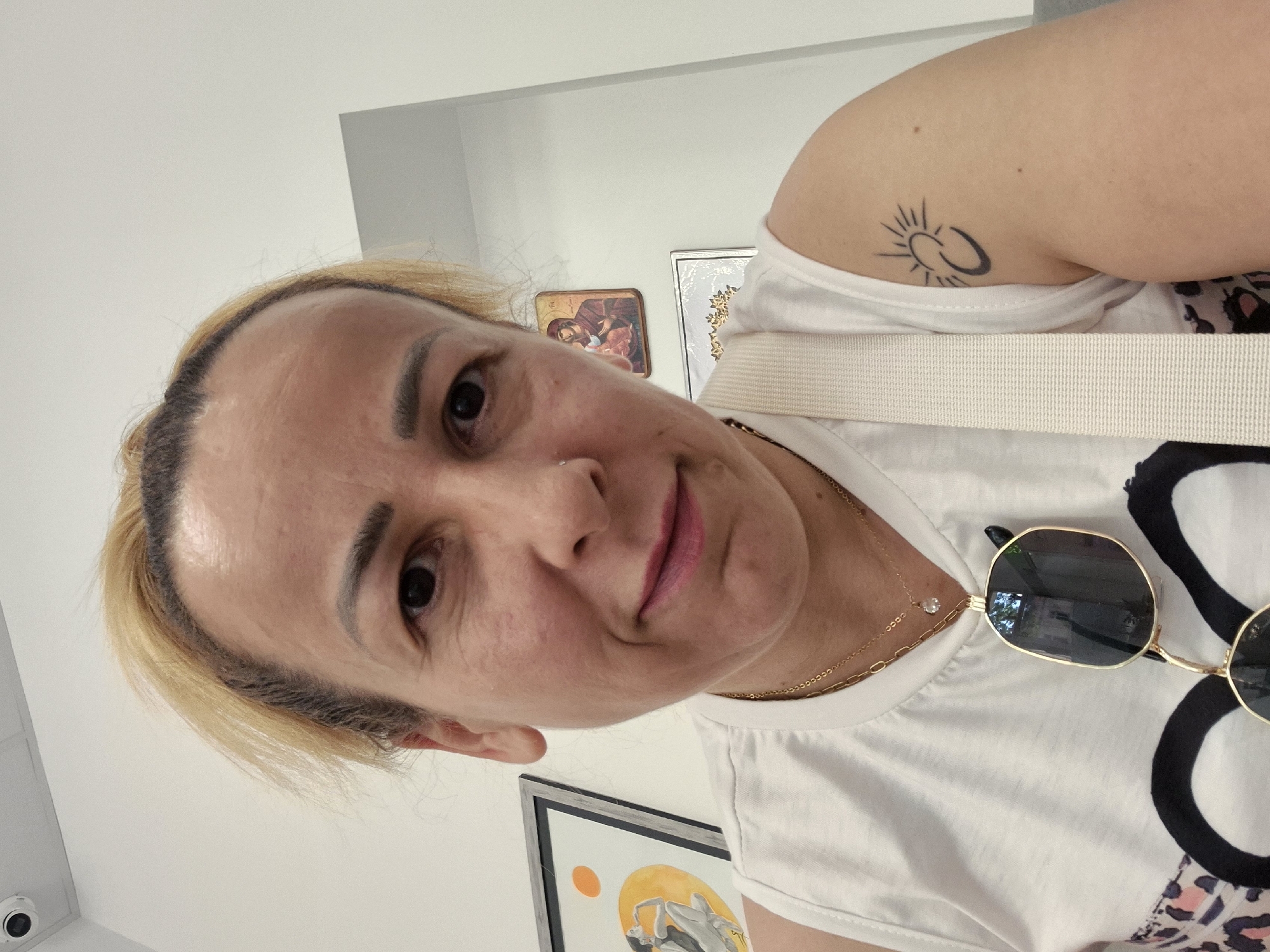 A selfie is taken by a person with blond hair, wearing a white tank top with a leopard print trim and a white strap across their chest. They are wearing two necklaces, one with a small diamond pendant. Sunglasses are hanging from the neckline of their shirt. The person has a small nose ring and a tattoo of a crescent moon and sun on their right arm. Behind them, on a white wall, are three framed pictures. The picture on the left is a colorful abstract with a sun and two figures. The middle picture is a religious icon of the Virgin Mary and child. The bottom picture is a white background with a gold decorative border. The ceiling has a white security camera visible.