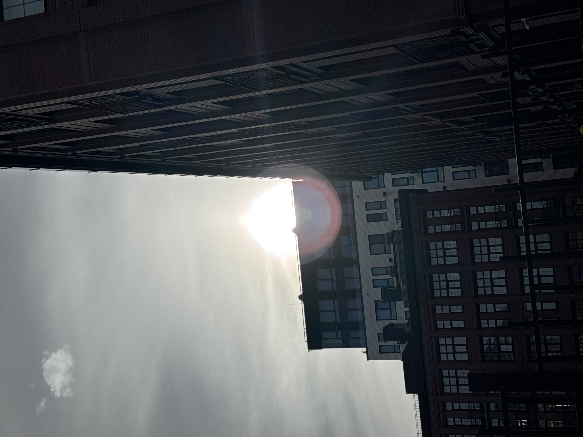 The image shows a view looking up between buildings in Moscow, Russia. The sun is visible, causing a glare. The sky is overcast. Several tall brick and concrete buildings are present, with windows. There is a small white cloud.