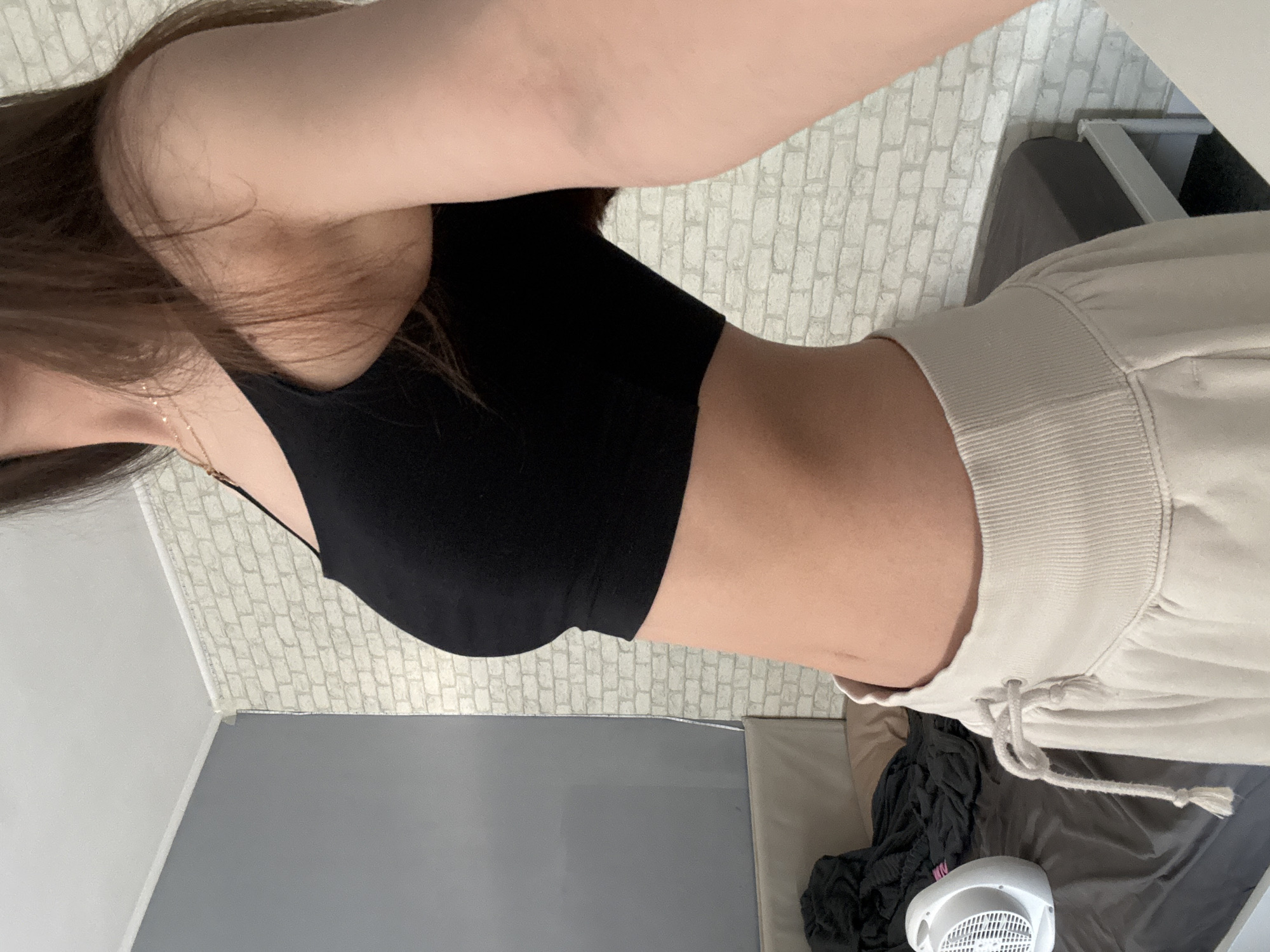 A young woman is posing for a selfie indoors. She is wearing a black crop top and light-colored sweatpants with a drawstring. A gold necklace is visible around her neck. Her hair is long and brown. Behind her is a textured white wallpaper and a gray wall. There is also a bed with gray and black bedding, and a white fan on the floor. There is no information in the image to indicate that this photo was taken in Sofia, Bulgaria.