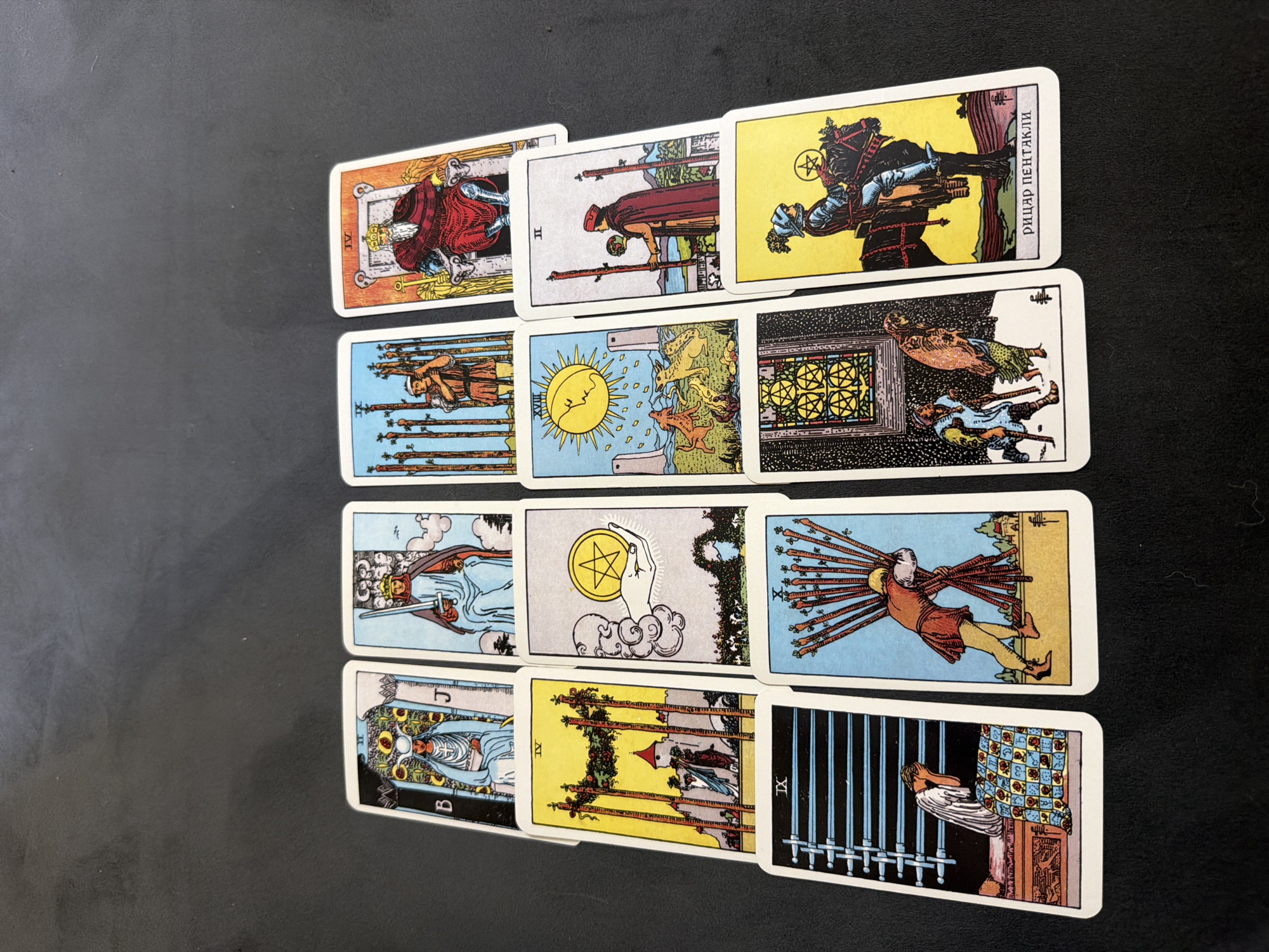 The image shows a spread of twelve Tarot cards arranged on a dark, textured surface. The cards are laid out in three rows. The top row has four cards: The Hierophant (labeled B and J), The Emperor (labeled IV), a card with a sun and dogs, and The Two of Wands (labeled II). The middle row has three cards: The Four of Wands (labeled IV), The Ace of Pentacles, and The Ten of Wands. The bottom row has five cards: The Nine of Swords (labeled IX), The Five of Pentacles, The Knight of Pentacles (labeled in Russian script), and another card that is partially obscured. The cards depict various symbolic figures and scenes, characteristic of Tarot card imagery. The lighting suggests an overhead light source, casting subtle shadows. The overall scene is static and appears to be a close-up photograph of the Tarot card spread.