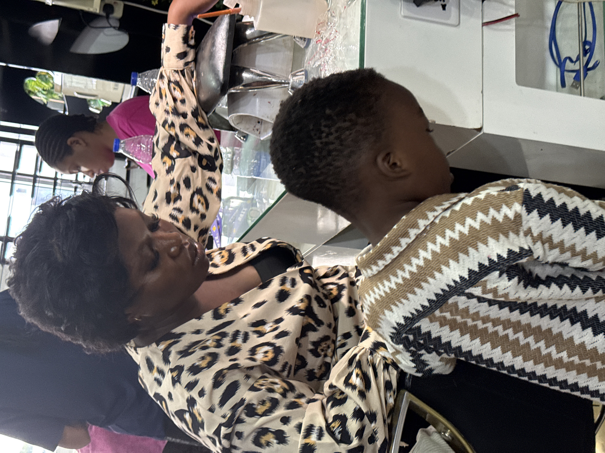 In an indoor setting in Lugbe, Nigeria, an adult woman with dark hair is seated, wearing a leopard-print blouse. A child with dark hair is beside her, wearing a zigzag-patterned shirt. The woman appears to be looking down at the child. Other people are visible in the background, and various objects are on a counter, including bottles and what appears to be a metal bowl. Electrical outlets and wires are also visible.