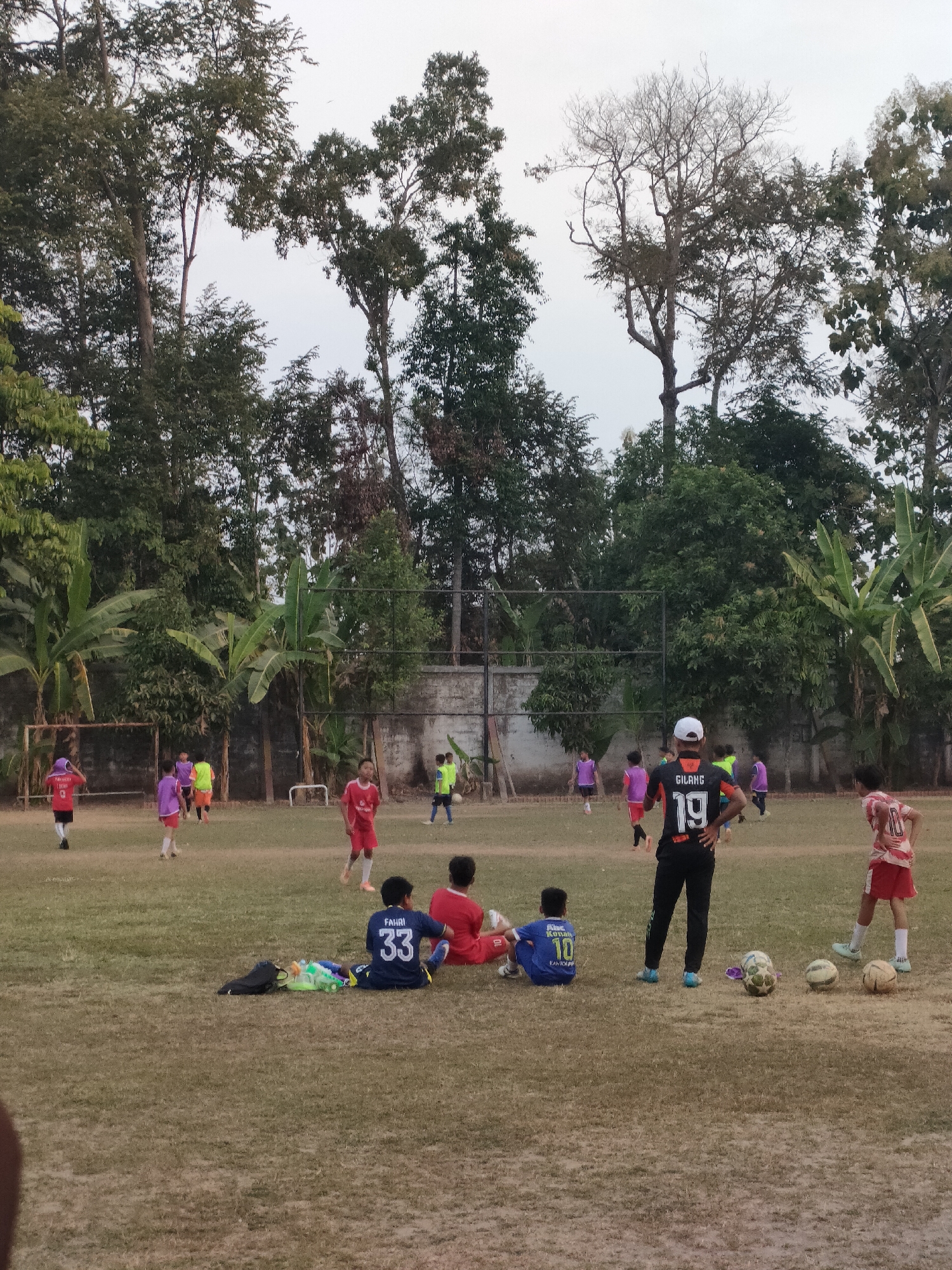 The image depicts a soccer field in Banguntapan, Indonesia, with a group of young males engaged in a soccer practice. The field is dry and grassy, with several trees and some fencing in the background. A group of boys are scattered across the field, some playing soccer, while others are resting. One boy in the foreground is sitting on the grass wearing a blue jersey with the number 33. Another boy in the background is wearing a black jersey with the number 19. Three soccer balls are visible on the ground. The sky is overcast, suggesting it might be late afternoon or early morning. The overall scene is one of casual outdoor activity.